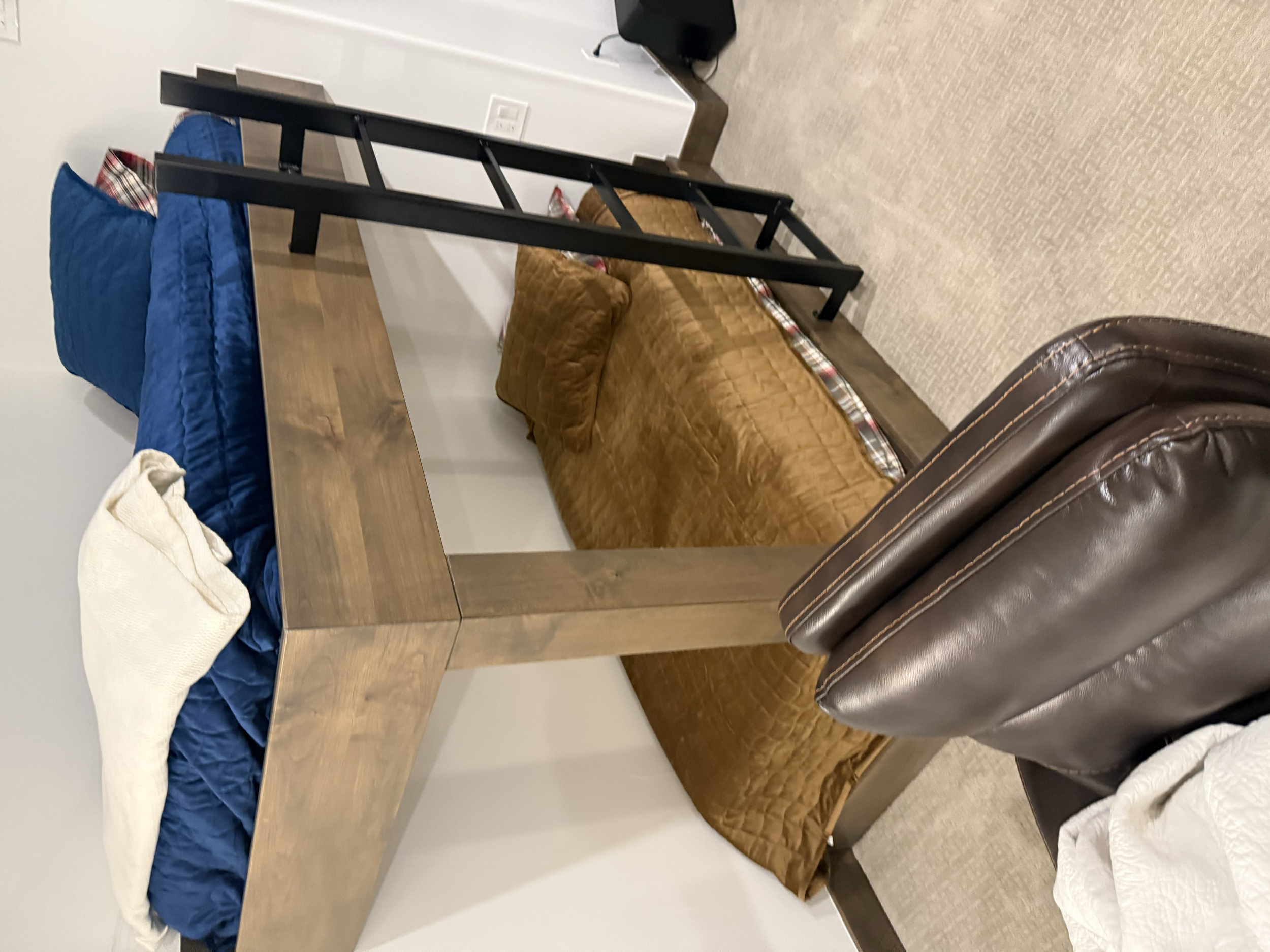 Room with a wooden table, a brown quilted couch with pillows, a black ladder, a leather chair, and a bed with a blue comforter and a white blanket.