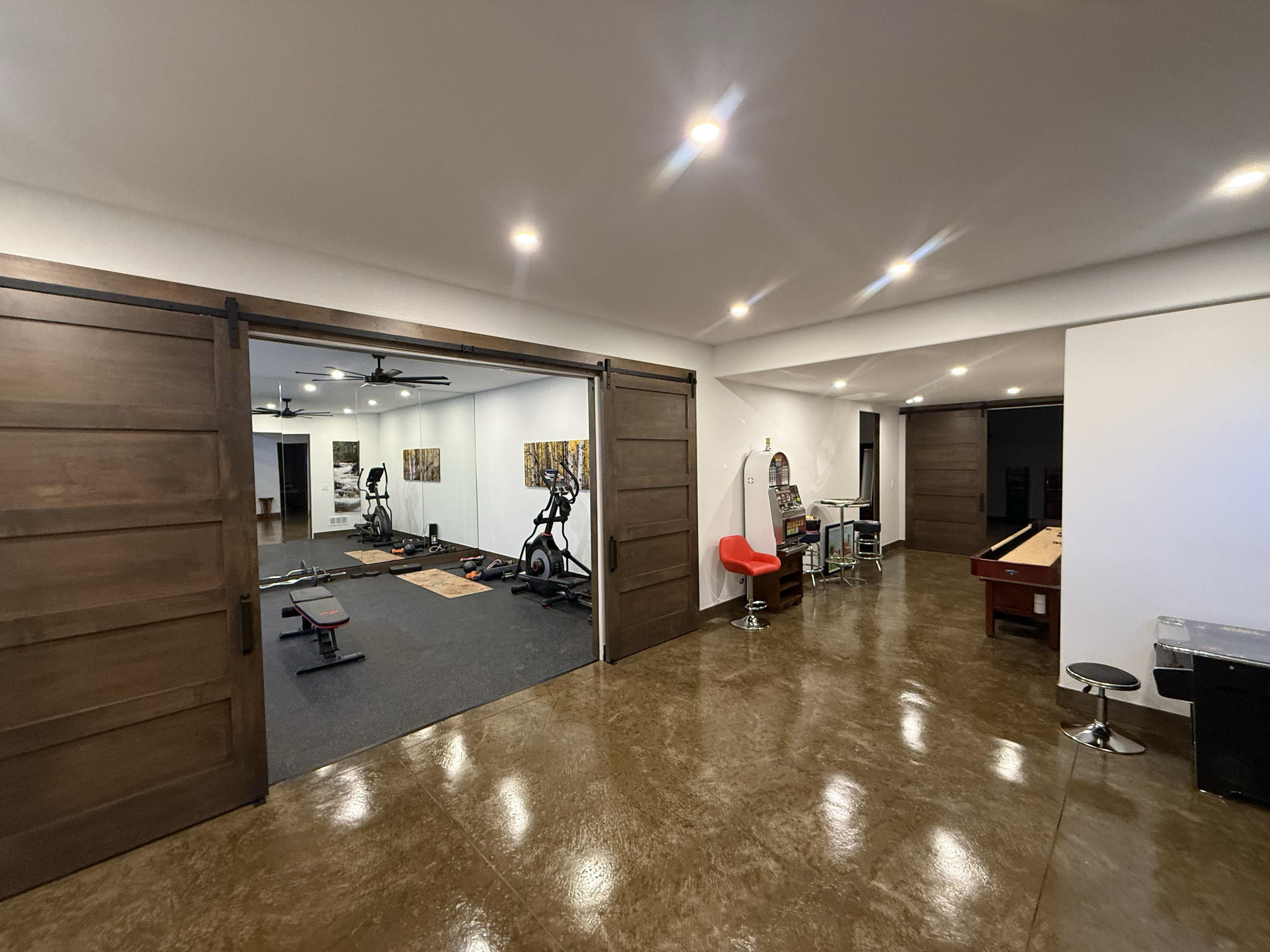 Interior view of a multi-purpose room with a wooden sliding barn door, equipped for exercise and entertainment, featuring gym equipment, arcade machine, and seating.