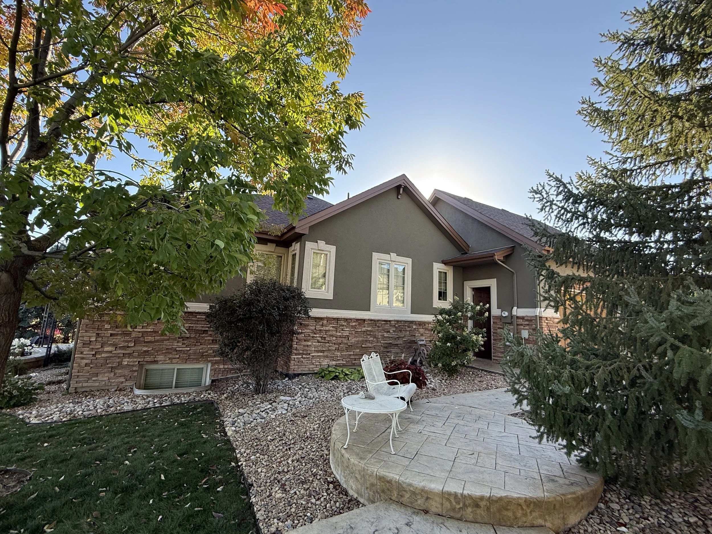 A residential home with a landscaped backyard, featuring a patio with white garden chairs, surrounded by green trees and shrubs, under a blue sky.