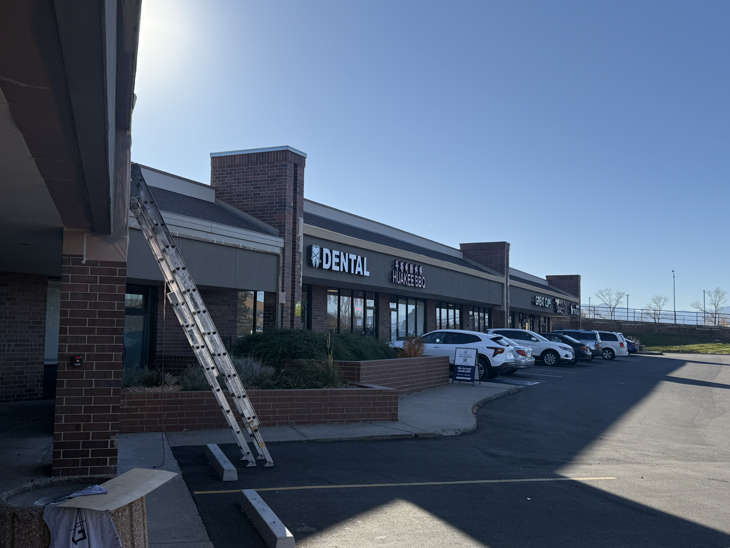 A strip mall showing stores including a dental office, Huakee BBQ, and Great Clips, with a parking lot in front and several parked cars under a clear blue sky.