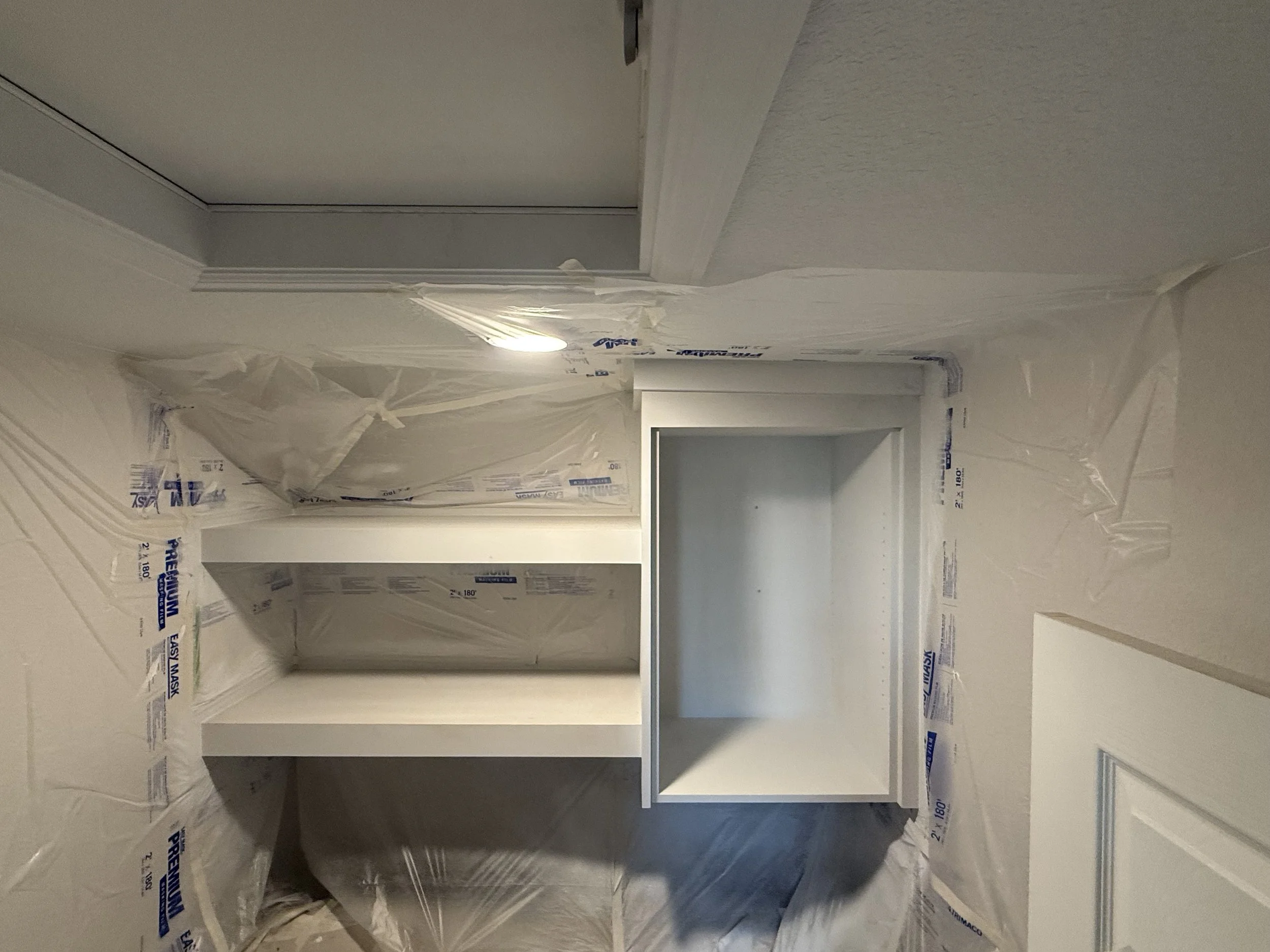 An interior room under construction with drywall and molding, with a recessed shelf built into the wall surrounded by painter's plastic sheeting.