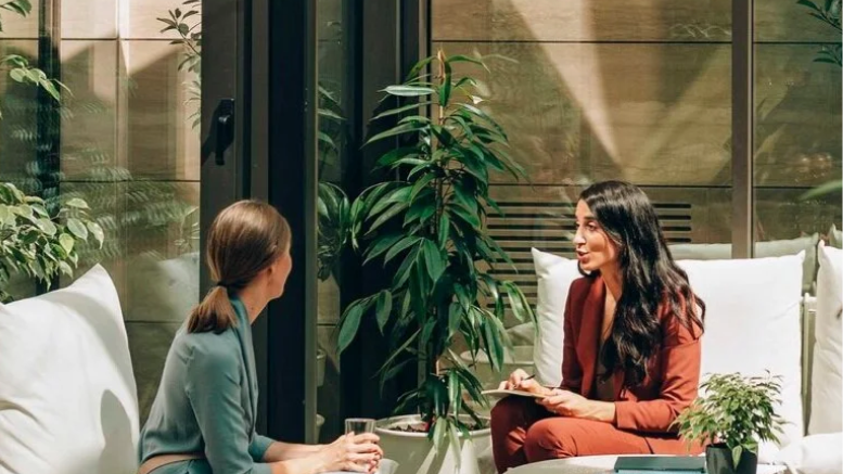 Two women engaging in a conversation in a cozy indoor setting with large glass windows, surrounded by plants and white cushions.