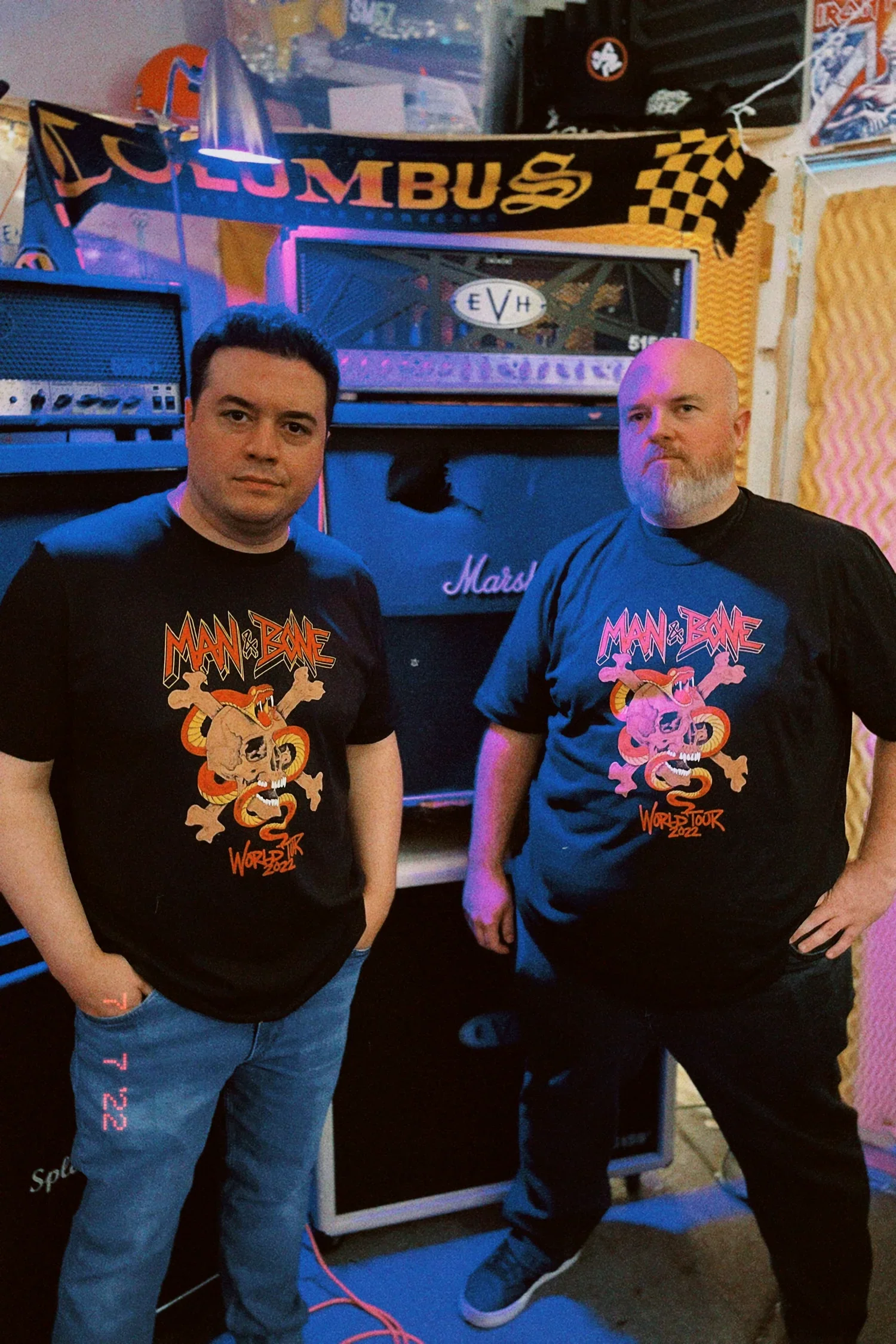 Two men standing in a music studio wearing matching black t-shirts with a logo and artwork that says "Man & Bone World Tour 2022". The background features musical equipment, posters, and foam soundproofing panels.