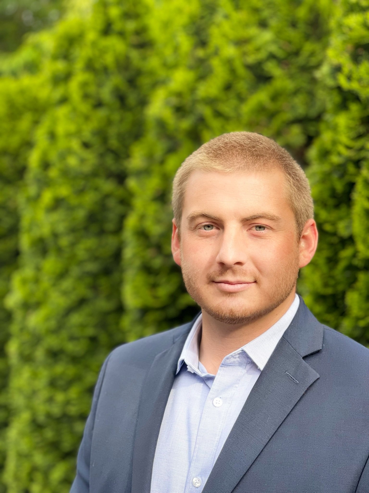 Cory Carlson, Founder of Constant Commercial Real Estate Inc and Constant Property Management LLC. Description: A young man in a blue suit and light blue shirt standing outdoors with a background of green foliage.