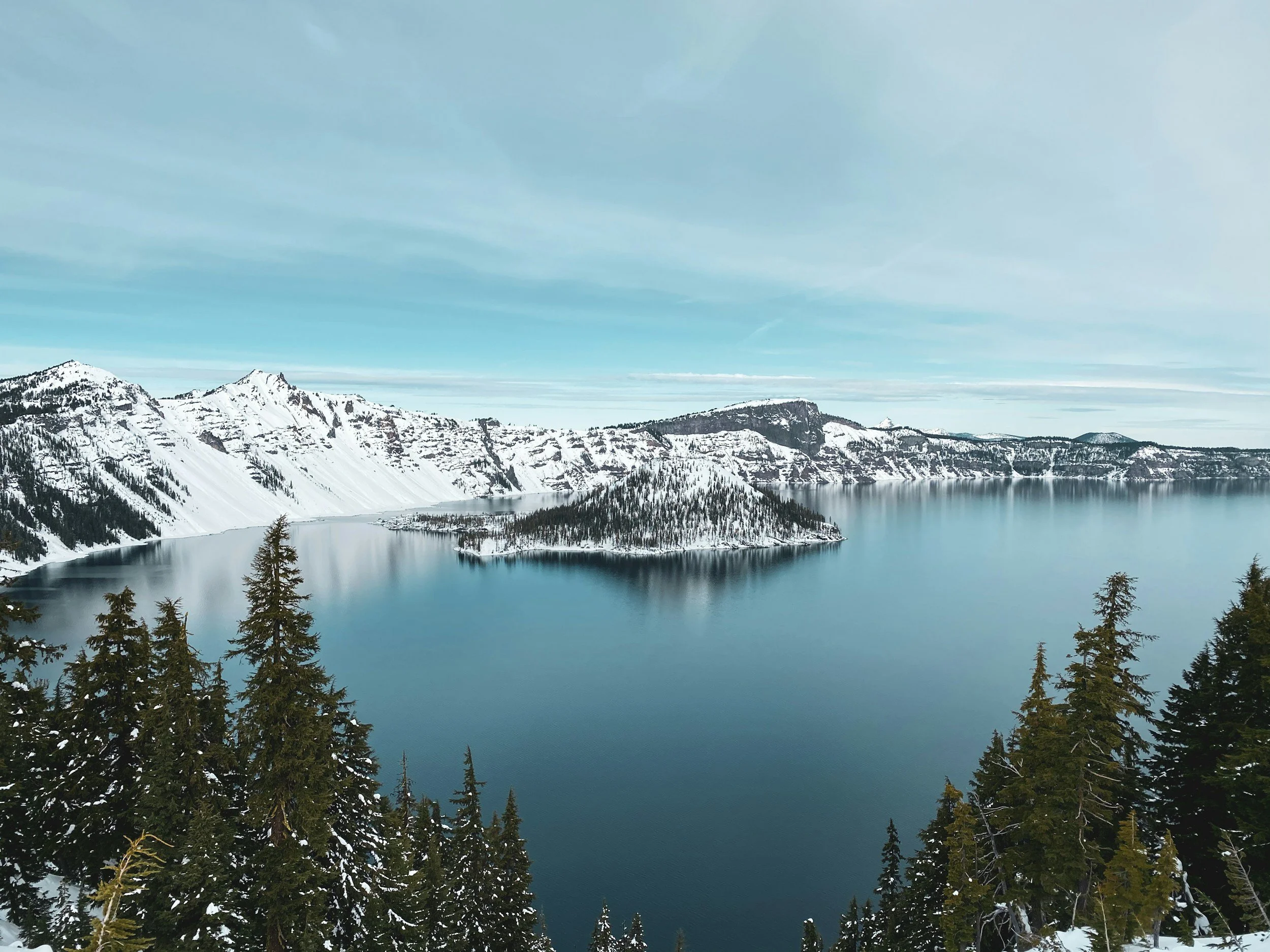 Crater Lake: A scenic view of a snow-capped mountain range surrounding a large, calm, blue lake, with tall evergreen trees in the foreground.