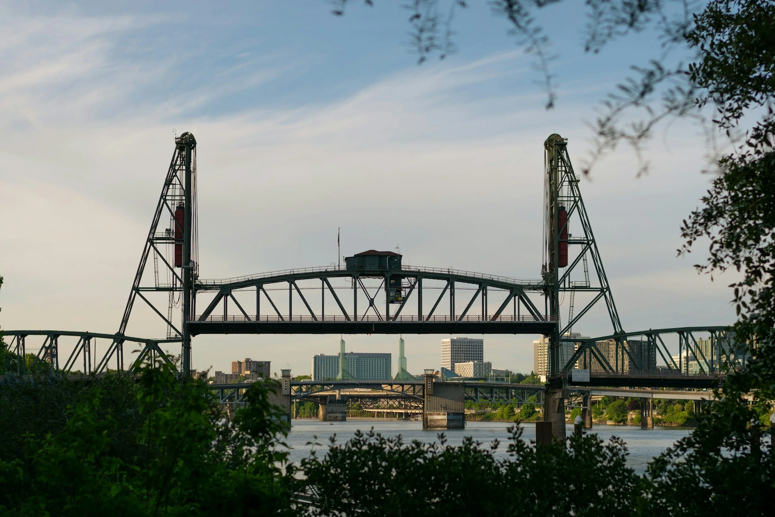 A large mechanical drawbridge over a river, with a city skyline in the background and trees in the foreground.