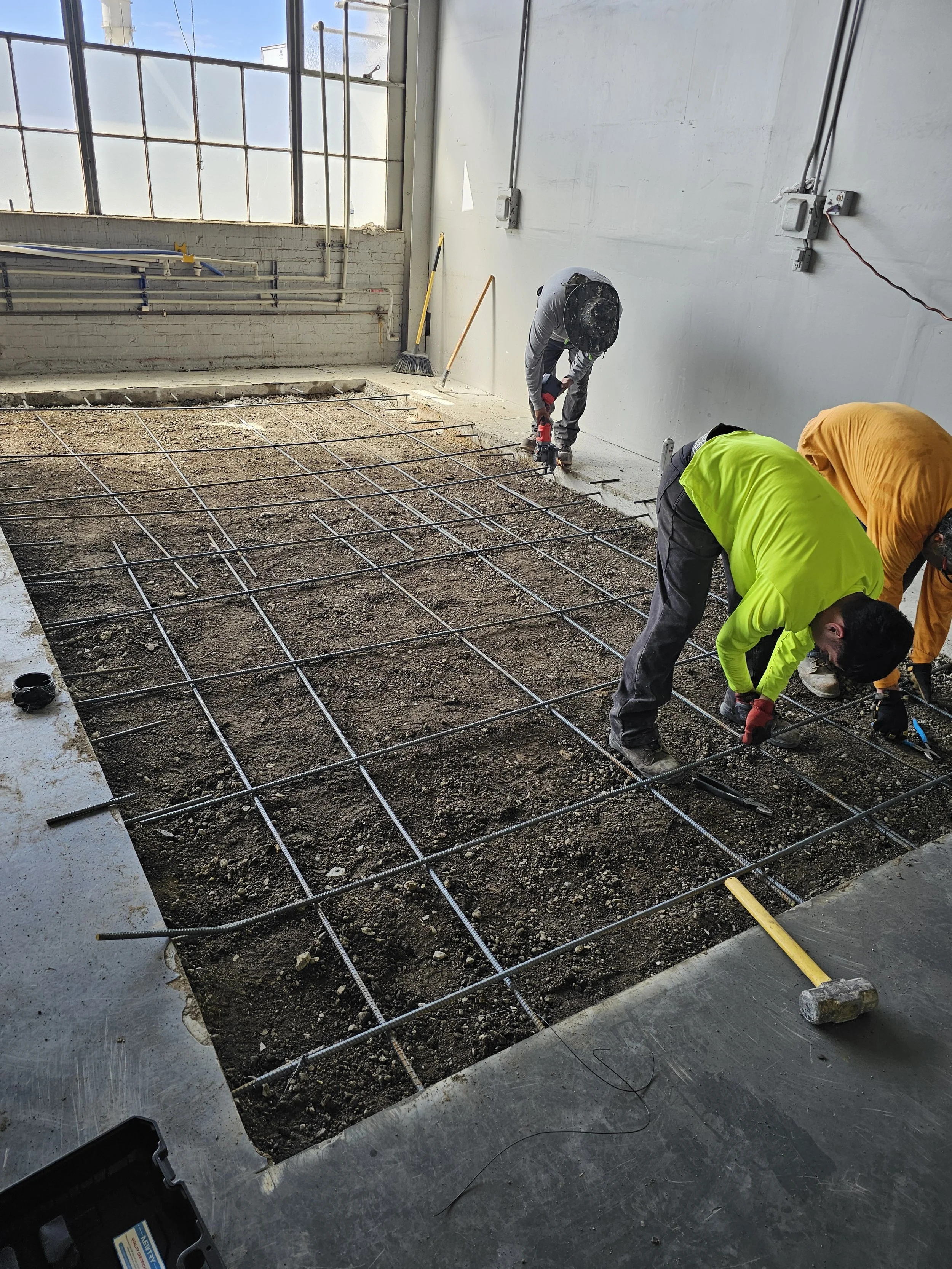 Construction workers installing rebar on a concrete floor inside a building, with tools and construction materials around.