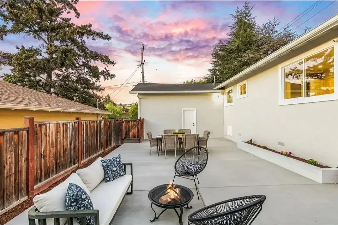 Backyard patio at sunset with outdoor seating, fire pit, wooden fence, and house with large windows.
