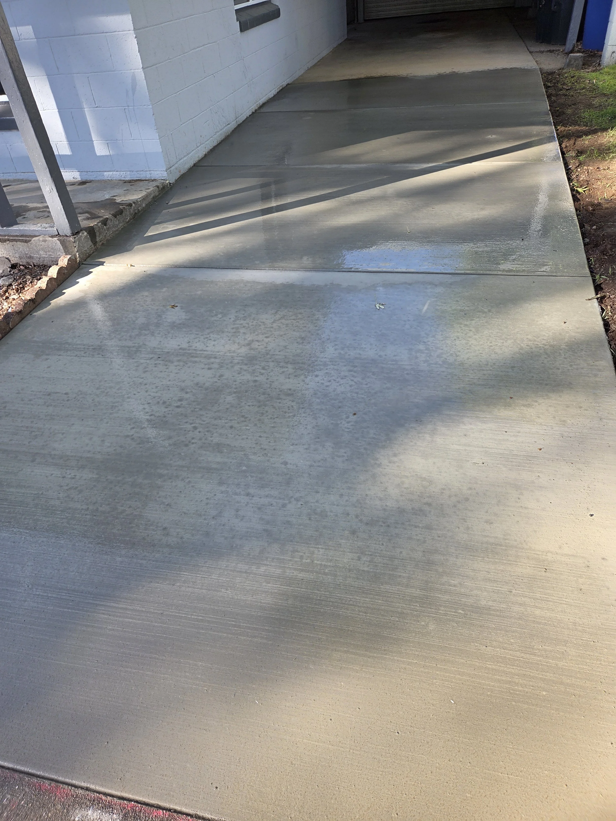 Freshly poured concrete sidewalk next to a white brick building with a metal railing on the left and a patch of grass on the right.
