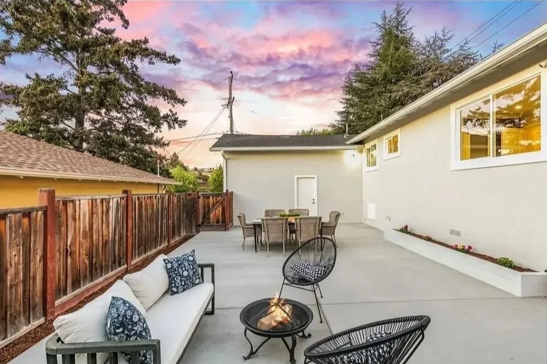 An outdoor patio area with a white bench, a fire pit, two black wire chairs, a dining table with six chairs, a wooden fence, and a white house with large windows during sunset.