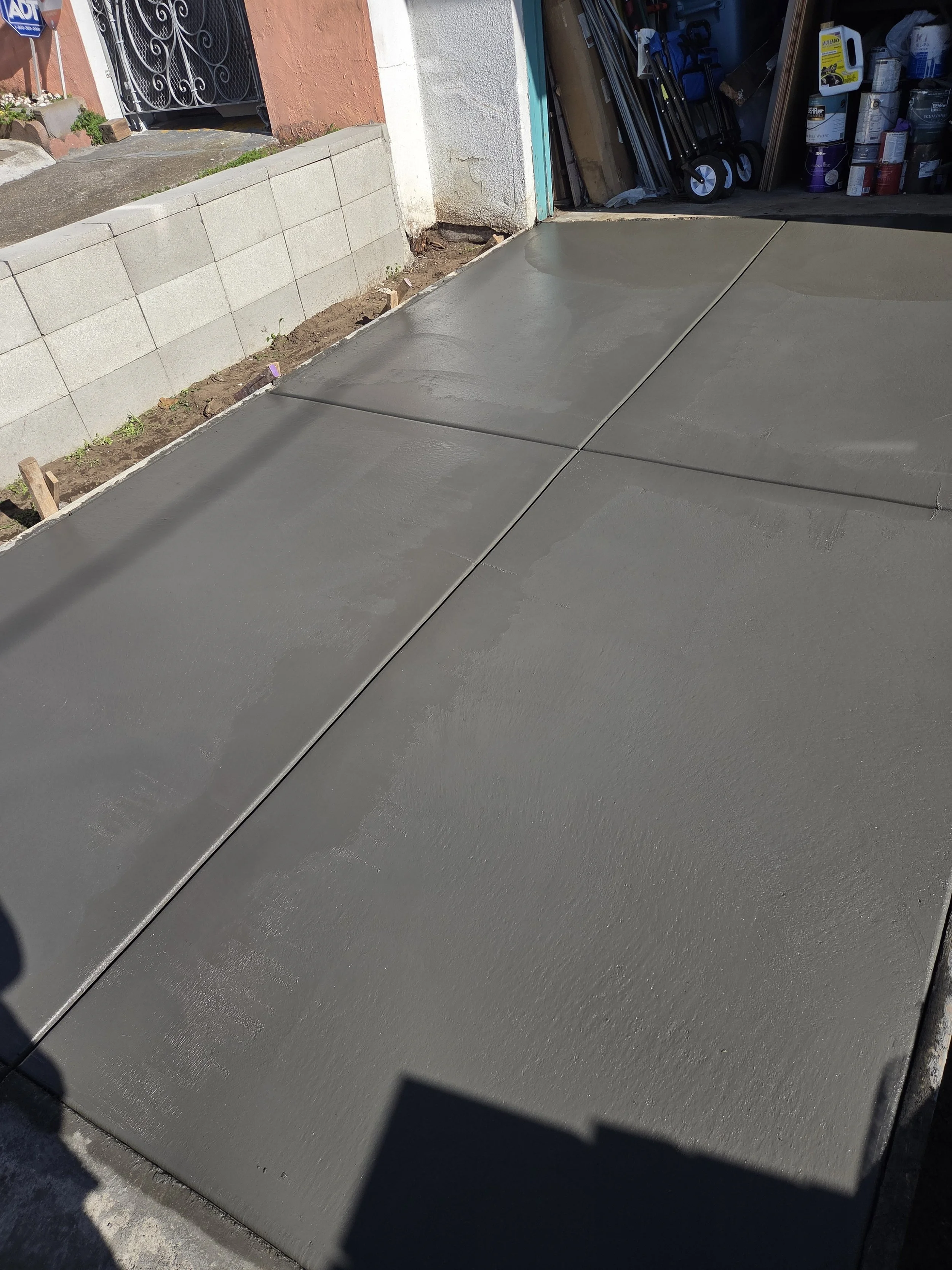 Freshly poured concrete sidewalk in front of a garage with gardening tools and supplies visible inside.