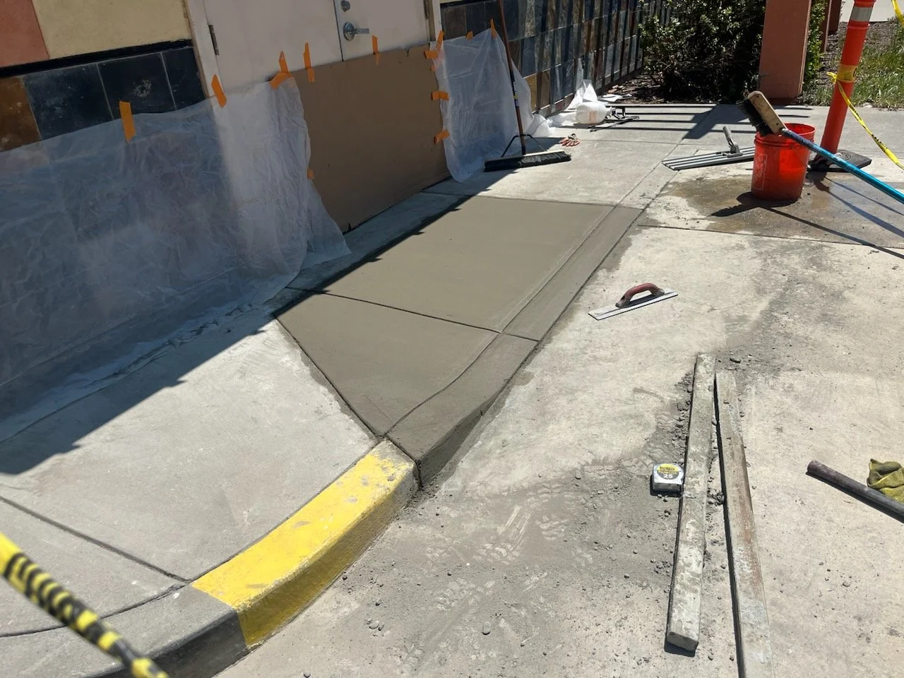 Concrete sidewalk being freshly poured and smoothed at a construction site, with tools and safety barriers nearby.