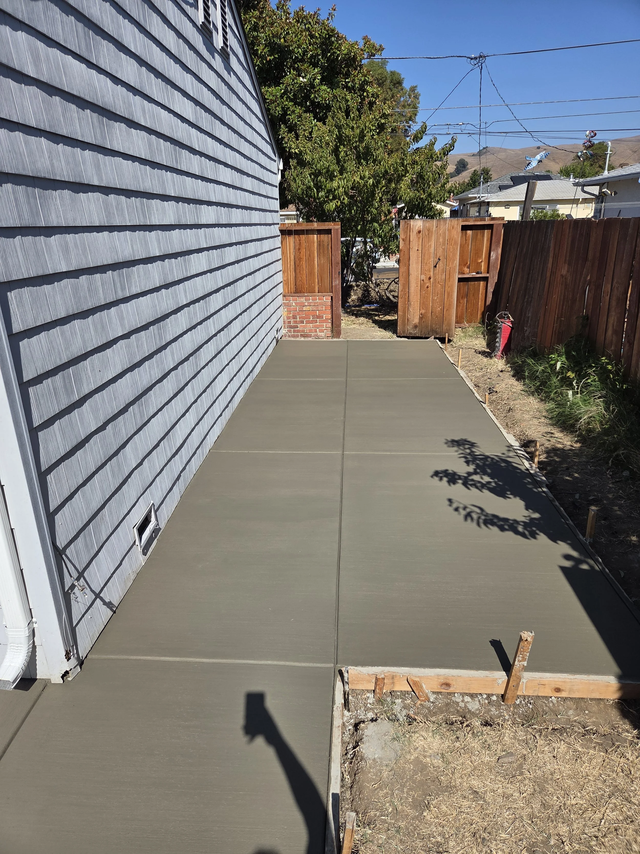 Freshly poured concrete patio with wooden framing, next to a blue house with siding, enclosed by a wooden fence, with some landscaping and tools nearby.