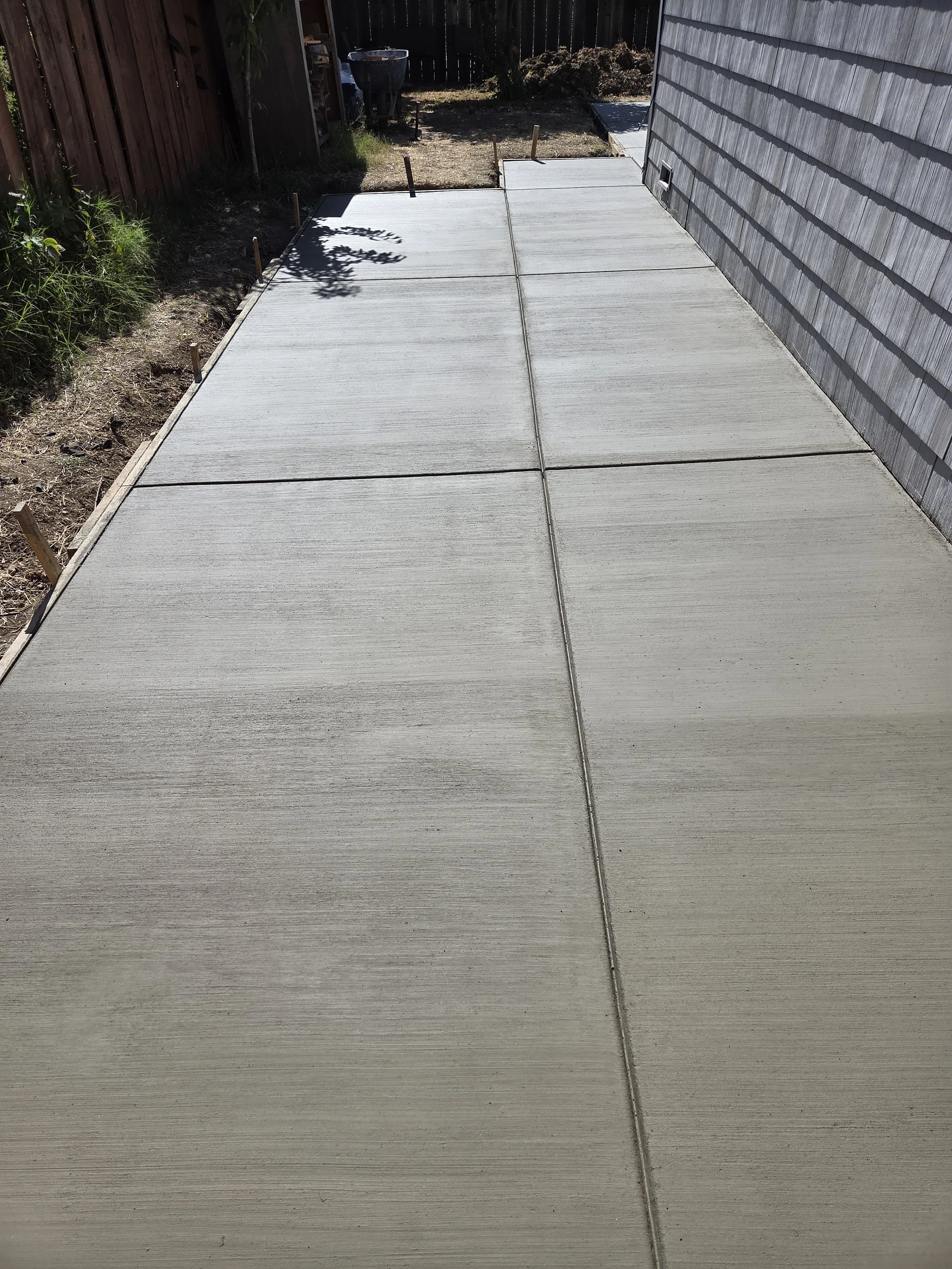 Newly poured concrete sidewalk next to a house with gray siding, with soil and plants on one side and a fence and garden area in the background.