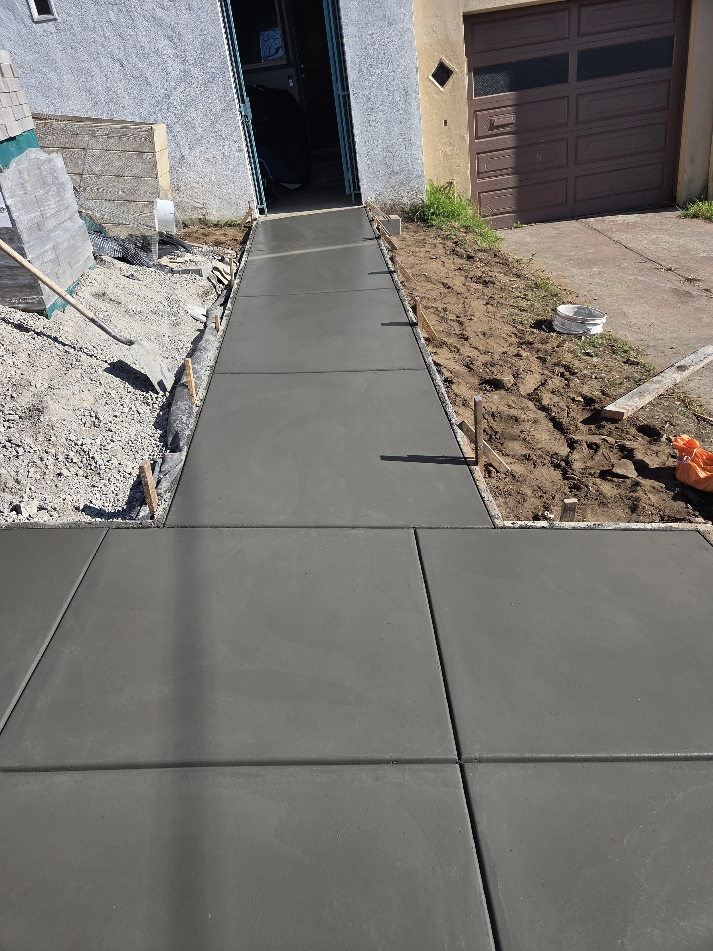 Newly poured concrete sidewalk leading to a house entrance with an open door, construction materials on the sides, and a partially finished yard.