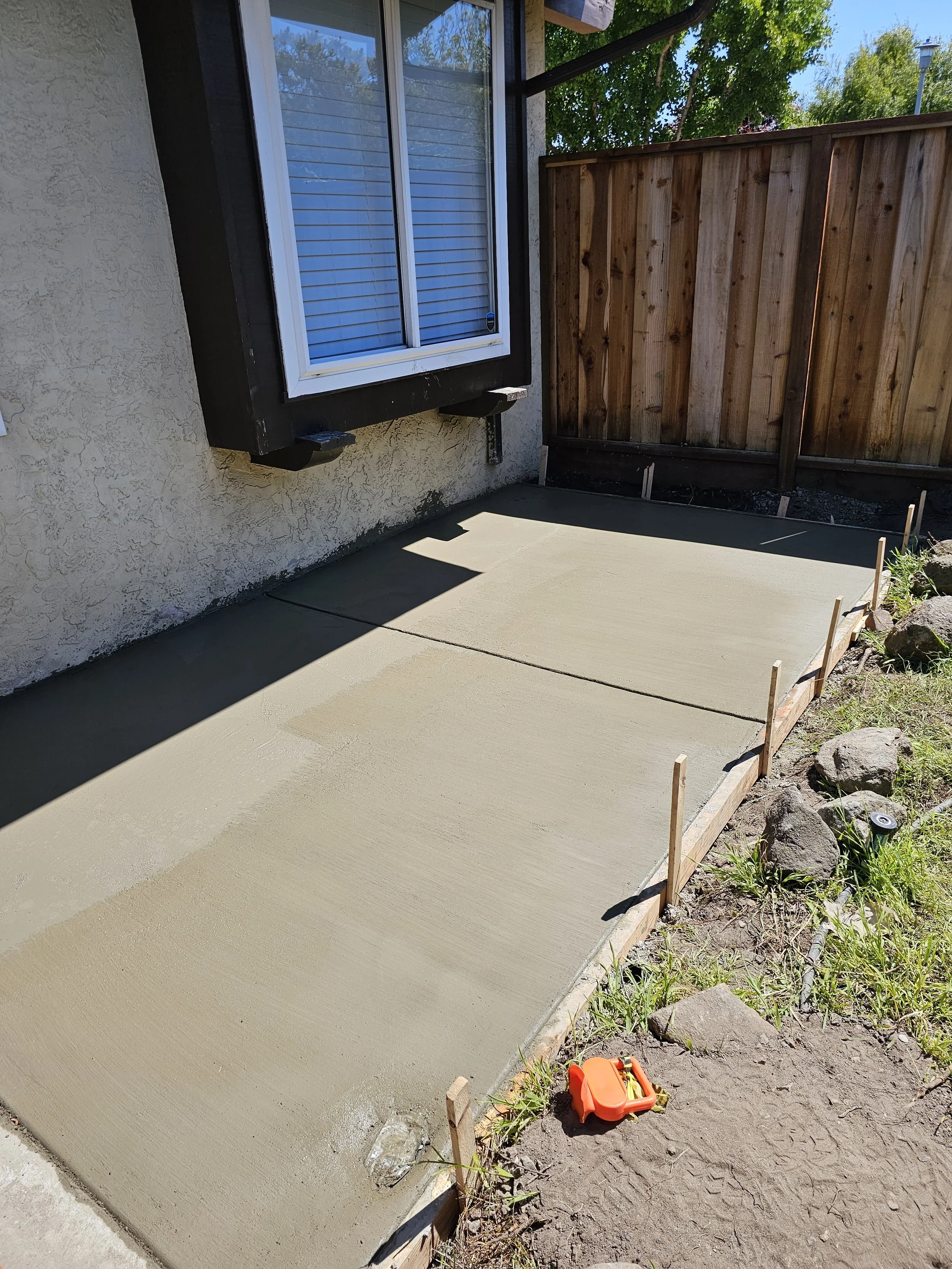 Freshly poured concrete on a small patio area next to a house with a window, surrounded by wooden stakes and a few rocks, with a yard and trees in the background.