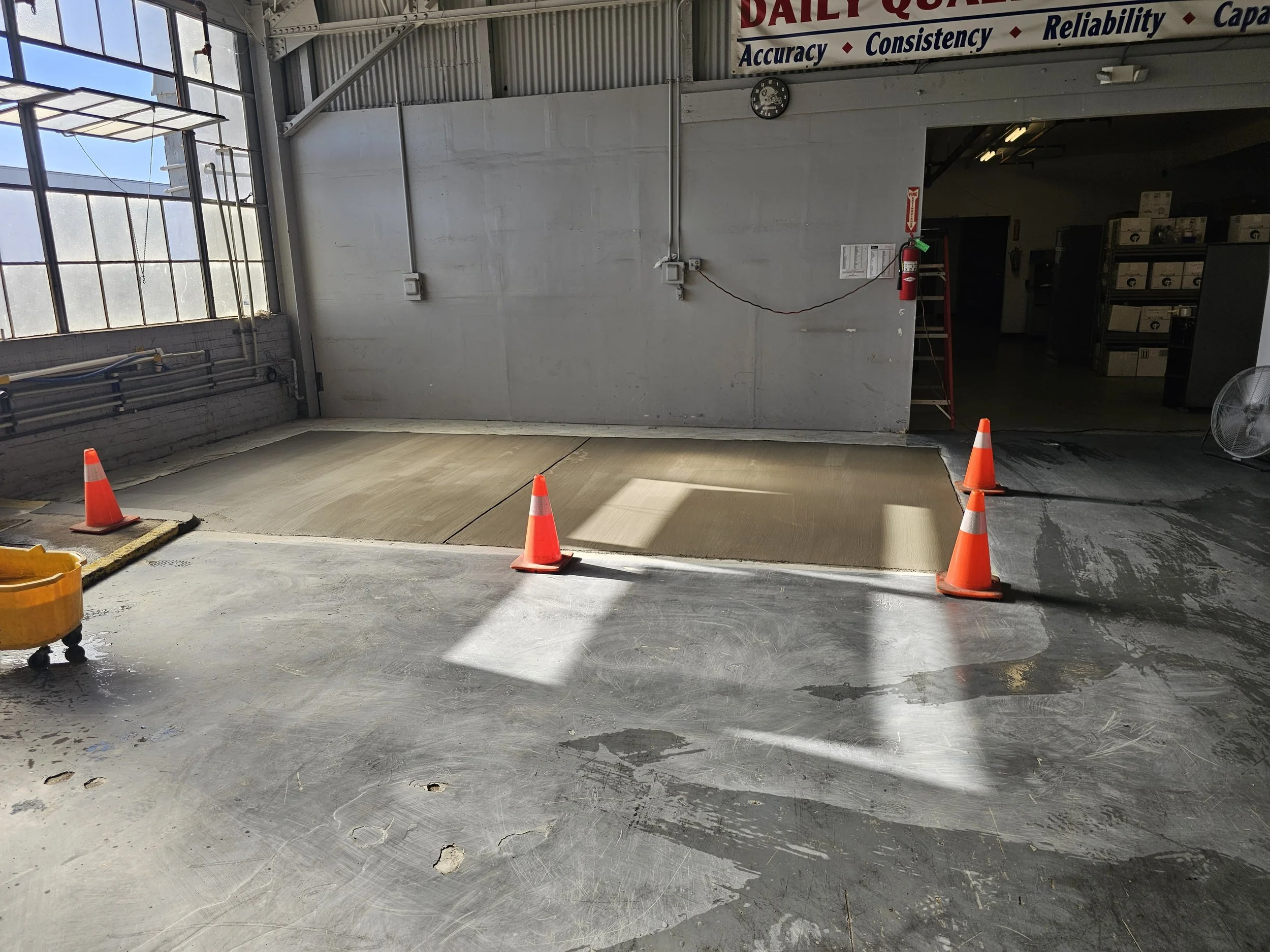 Construction area inside a building with a concrete floor, four orange traffic cones, and a new concrete pad in the center.