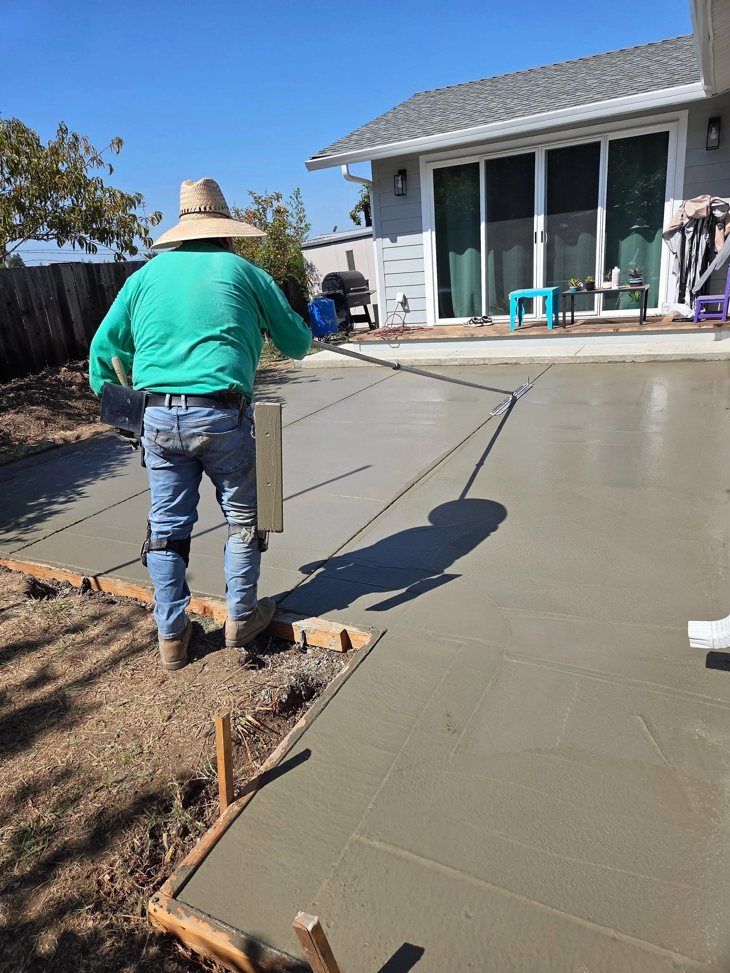A person wearing a straw hat and green shirt is pouring liquid concrete on a freshly poured concrete patio next to a house with large sliding glass doors, with a clear blue sky above.