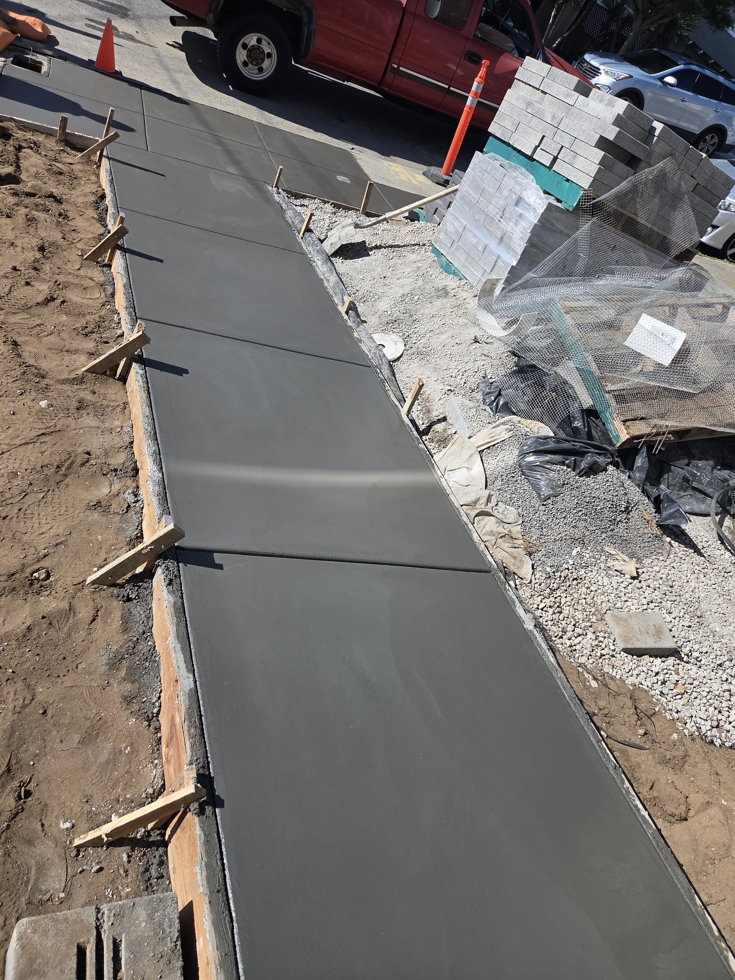 Freshly poured concrete sidewalk under construction with wooden forms and rebar, construction materials and vehicles in the background.
