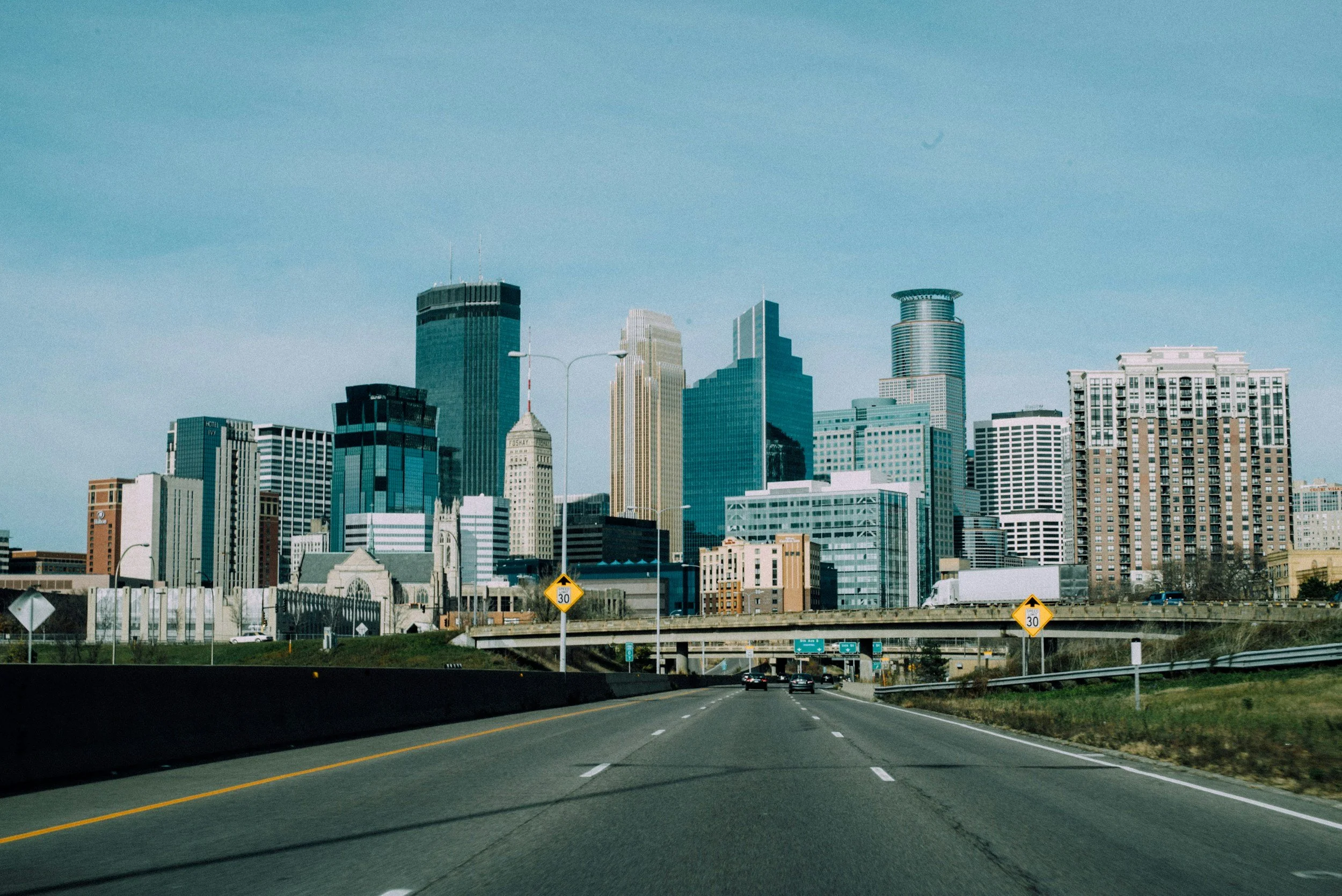 View of a city skyline with tall modern buildings, taken from a highway with a few cars, under a blue partly cloudy sky.