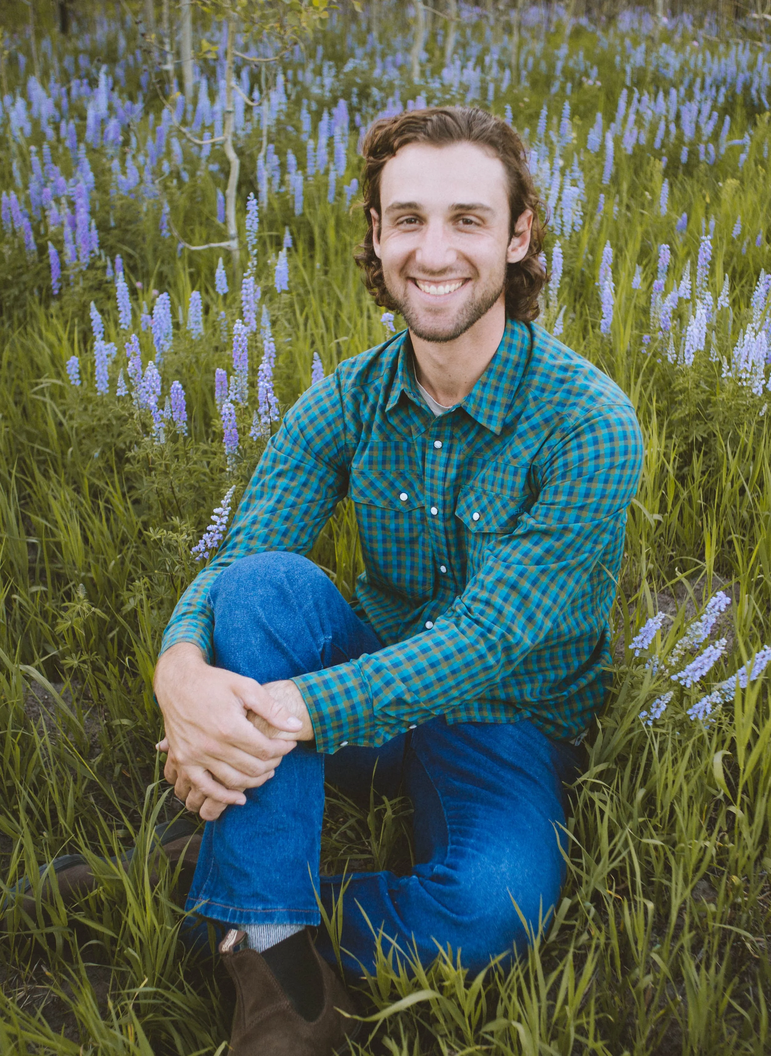 A young man with brown hair and a beard sitting in a field of purple flowers, smiling at the camera. He is wearing a green and blue checkered shirt and blue jeans.