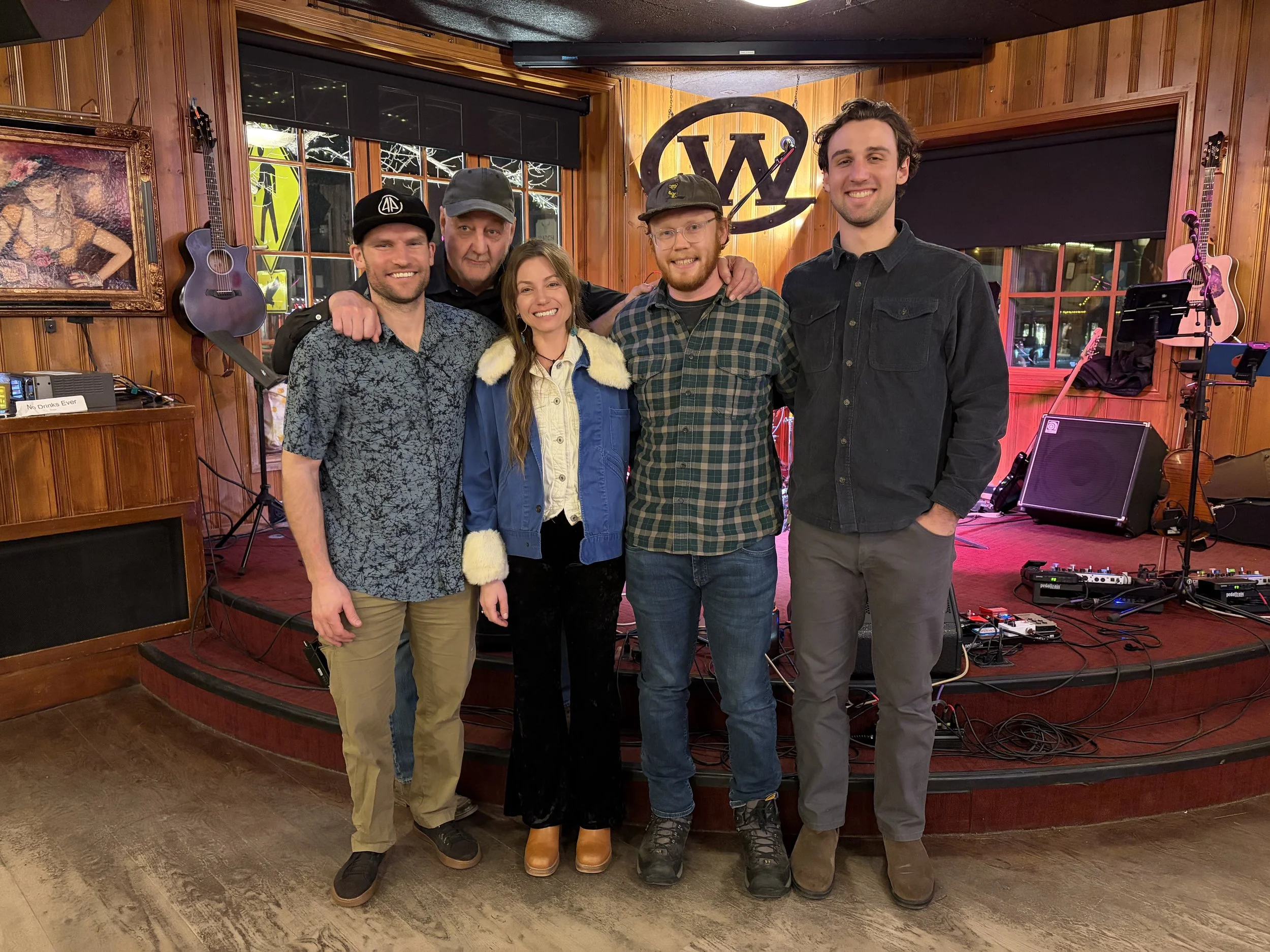 A group of five people standing on a stage in a wooden room, smiling at the camera. The stage has musical instruments and equipment, with a large W logo behind them. The background includes guitars and paintings, with large windows showing the outsid