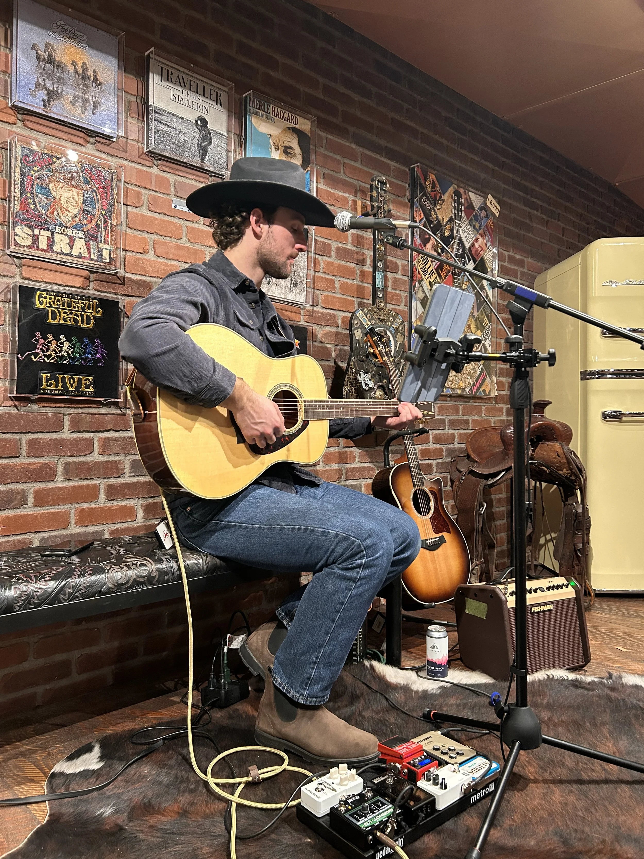 A man playing an acoustic guitar in a room decorated with vintage music record covers, standing in front of a brick wall, with a microphone, guitar pedals, and an amplifier nearby.