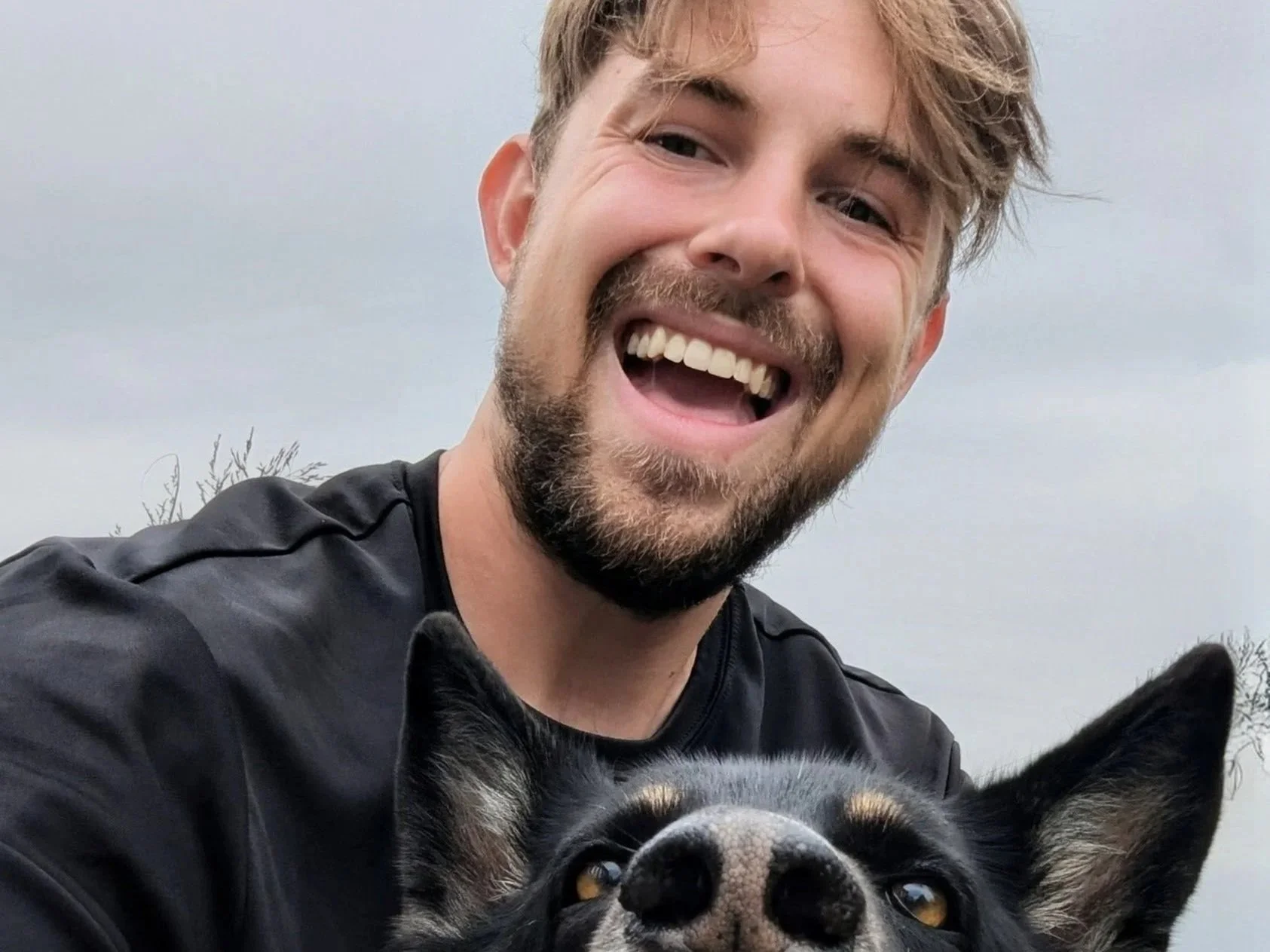 A smiling man with brown hair, beard, and mustache taking a selfie with his black dog outdoors against a cloudy sky.