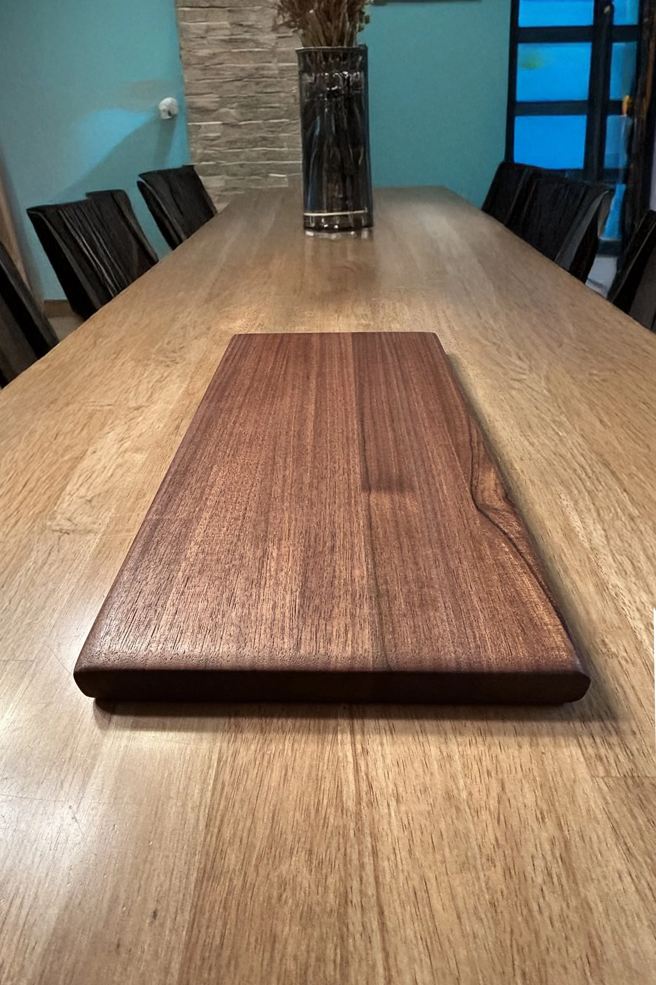 A long wooden dining table with a rectangular wooden serving board placed in the center. There are black chairs around the table, a tall glass vase with dried flowers, and a blue wall with a window in the background.