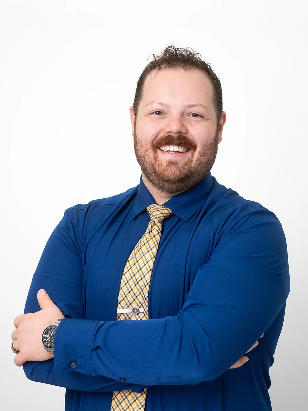 A smiling man in a blue dress shirt with a yellow plaid tie, crossed arms, and a wristwatch, standing against a plain white background.