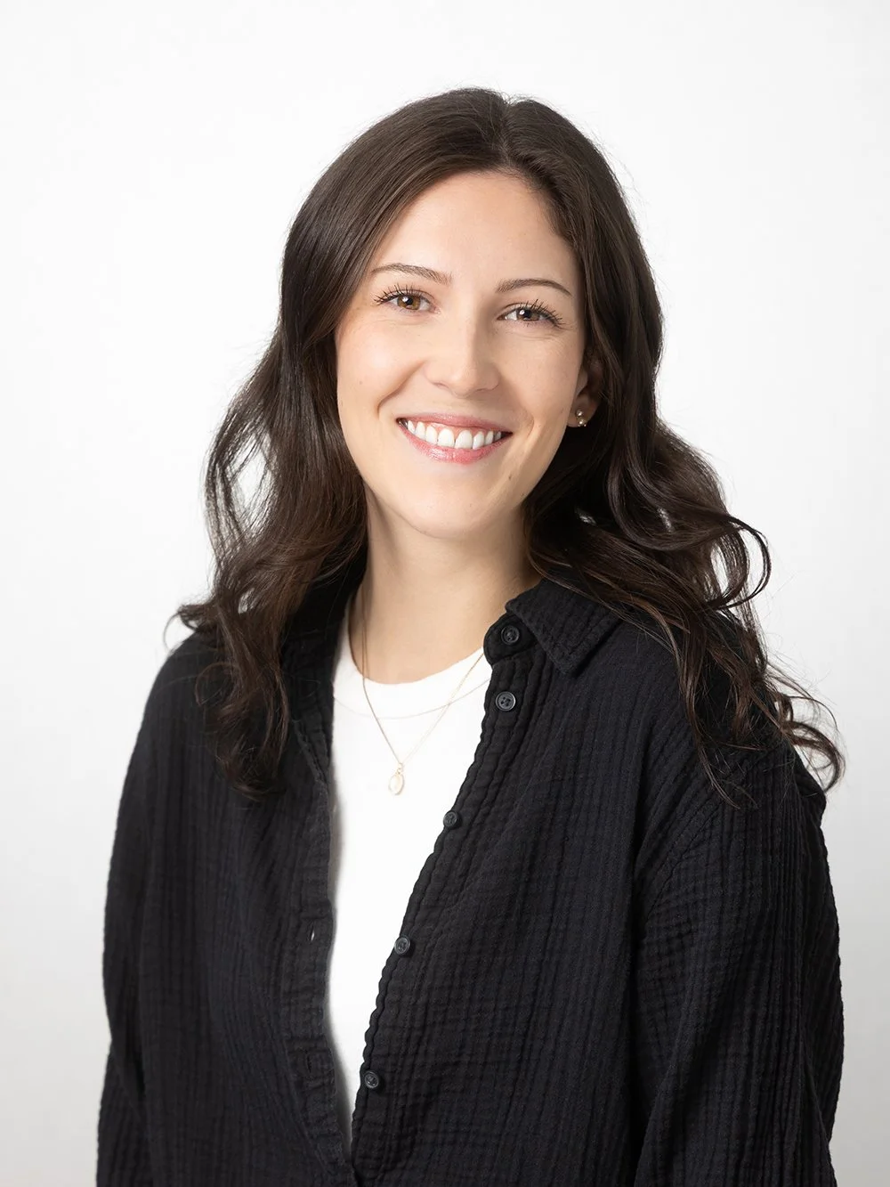 A young woman with long dark hair, smiling, wearing a black button-up shirt and a necklace, against a plain white background.