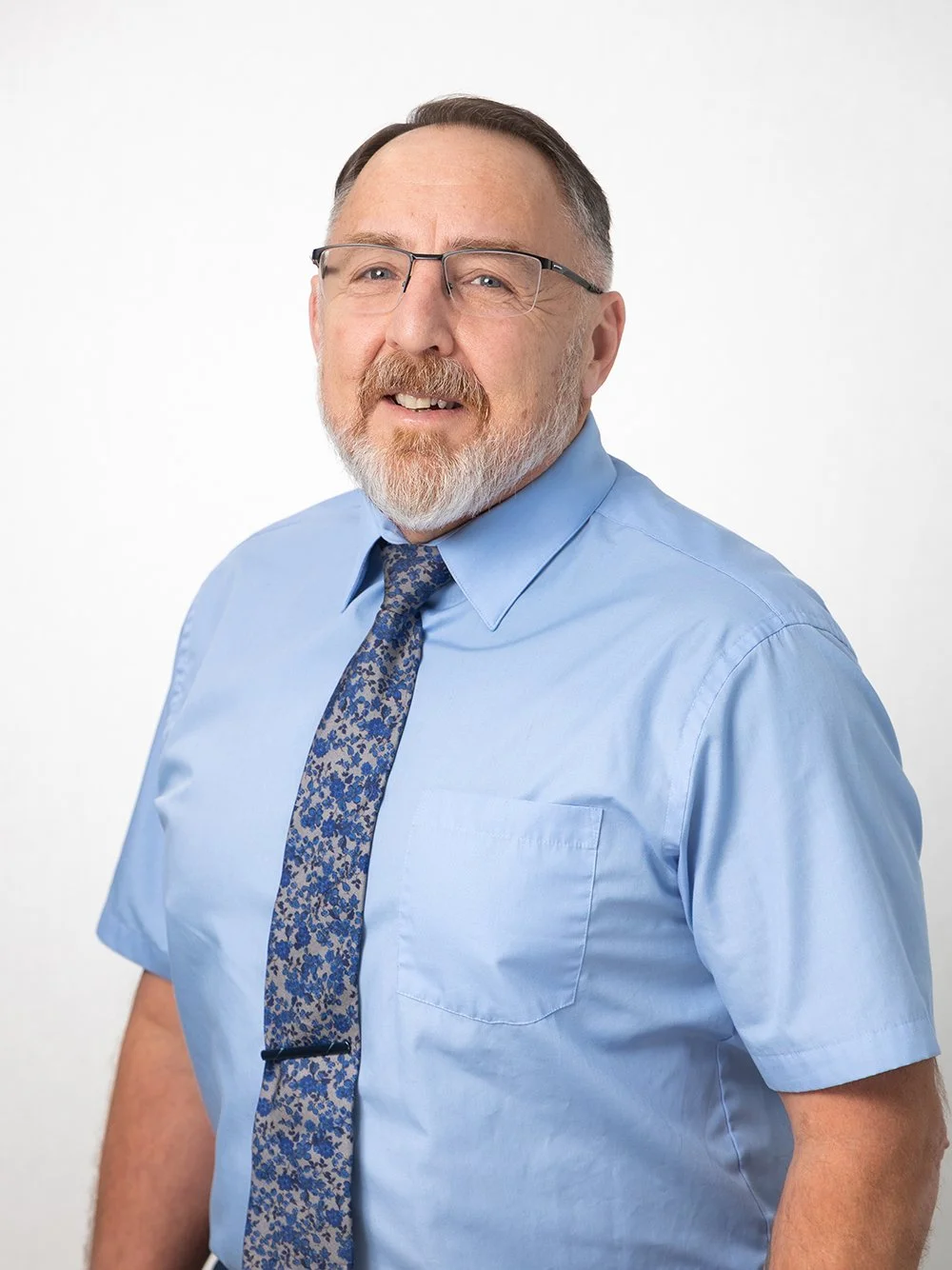 A middle-aged man with glasses, a beard, and a mustache wearing a light blue shirt and a patterned blue tie, smiling against a plain white background.