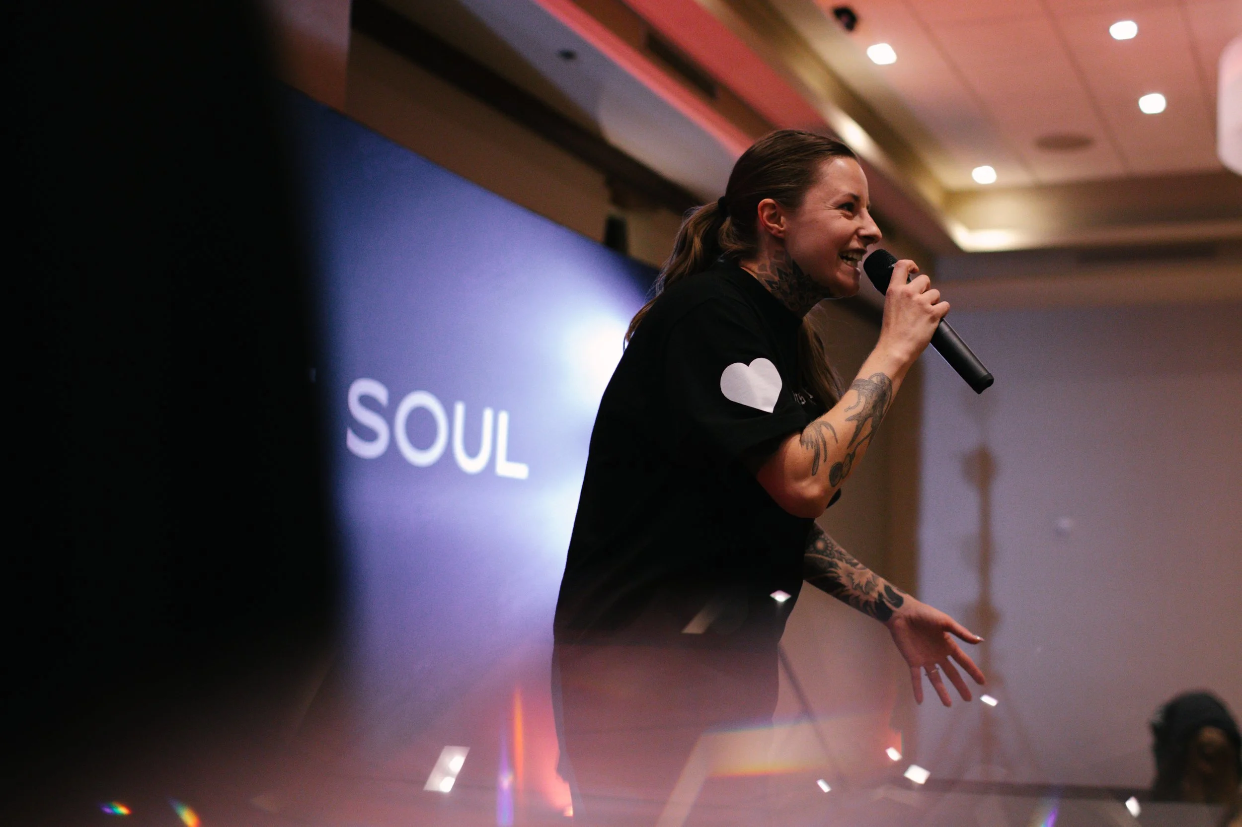 A person with tattoos on their arms, smiling and speaking into a microphone at an event. They are wearing a black shirt with a white heart on the sleeve, and behind them is a screen with the word 'SOUL' illuminated.