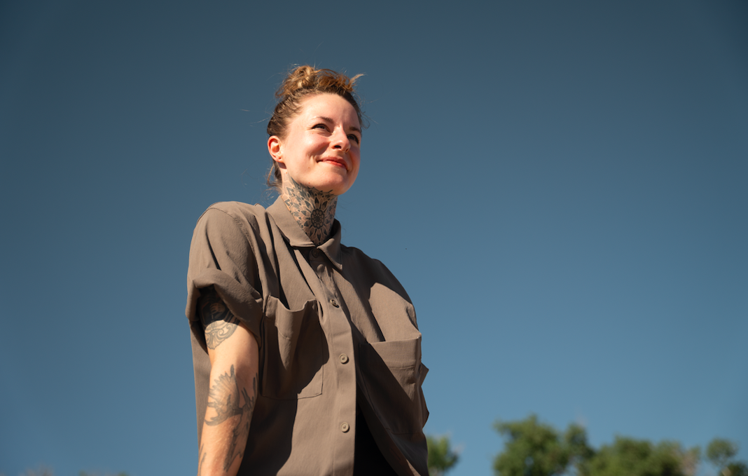 A woman with tattoos on her arm and neck standing outdoors against a blue sky, smiling.
