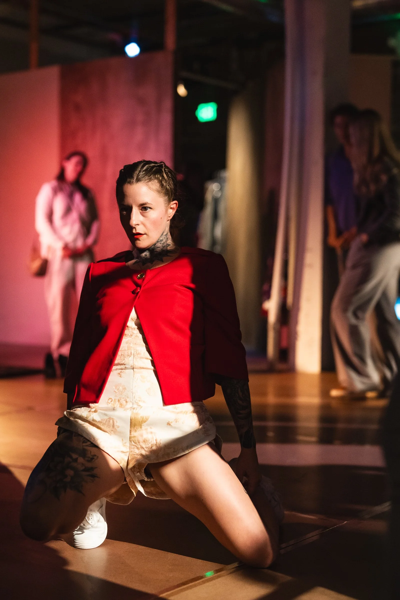 A person in a red jacket and beige shorts with tattoos on her arms and legs, kneeling on the floor, with a serious expression. In the background, other people are standing and watching in a dimly lit indoor space.