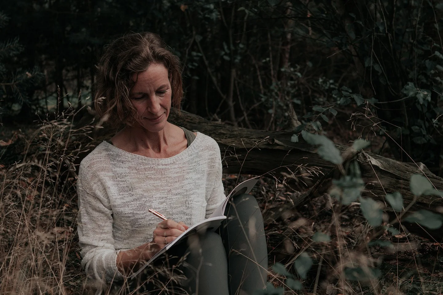 A woman in a white sweater sitting outdoors among trees and dry grass, writing in a notebook with a pencil.