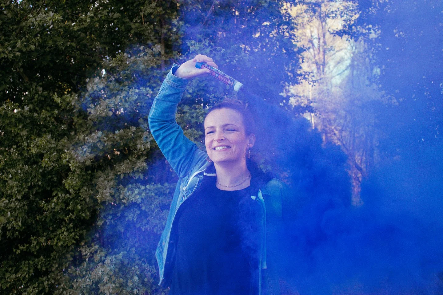 A woman outdoors in front of trees, holding a spray can and smiling, with blue spray paint in the air around her.