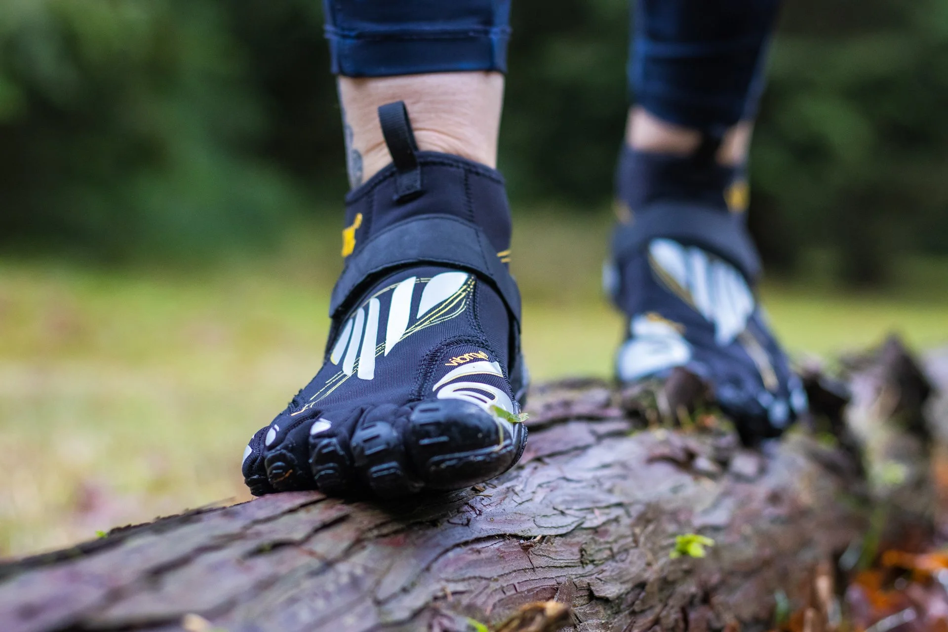 Close-up of a person wearing black and yellow protective biking shoes balancing on a fallen tree trunk in a forest.
