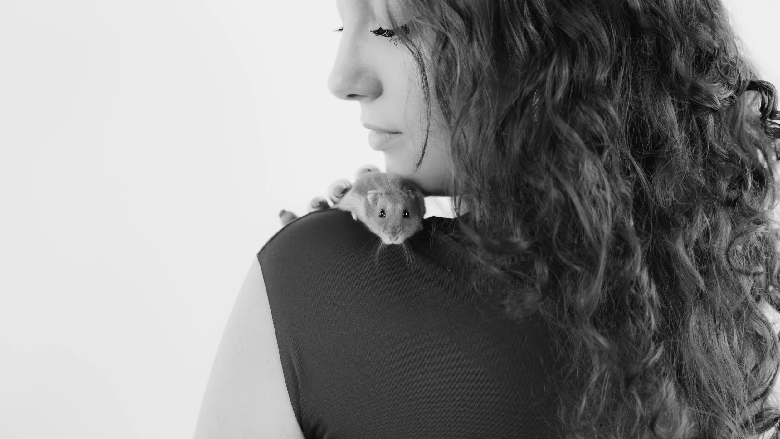 A woman with curly hair looking down at a small hamster perched on her shoulder, against a plain light background.