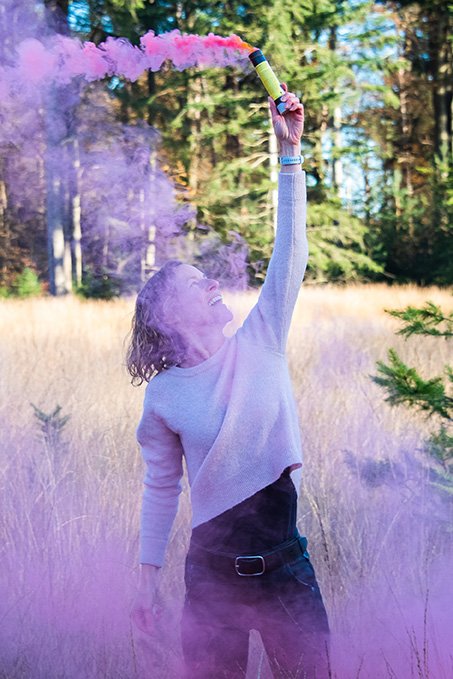 A woman in a light sweater and dark pants holding a smoke bomb in the air, emitting colorful pink smoke, standing in a meadow with trees in the background.