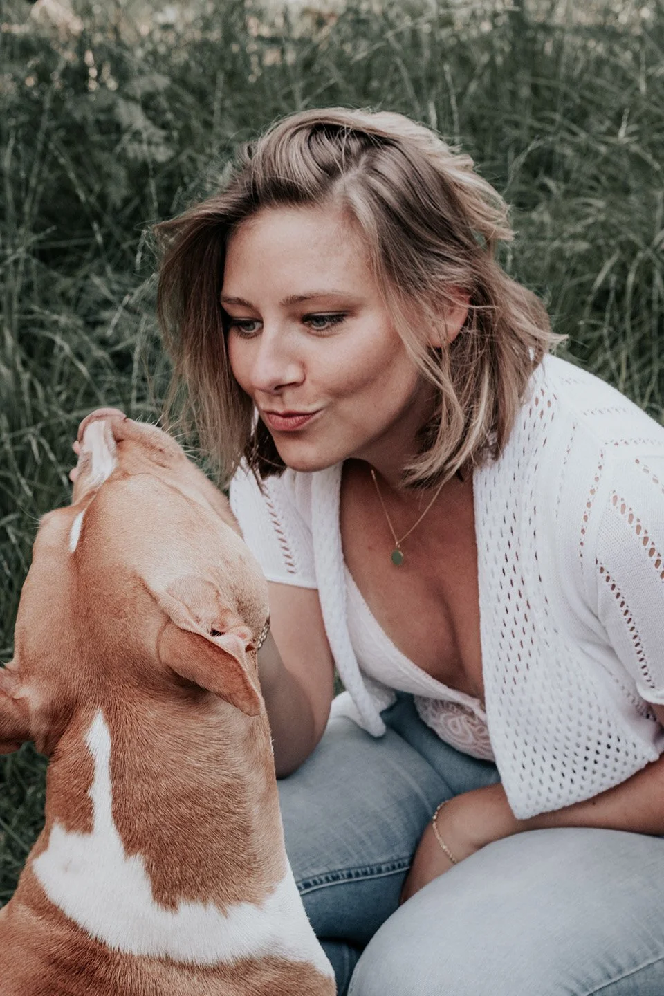 A woman with shoulder-length blonde hair and a white crochet top is outdoors, leaning in towards a tan and white dog, appearing to be blowing a kiss or making a playful face at the dog, with greenery in the background.