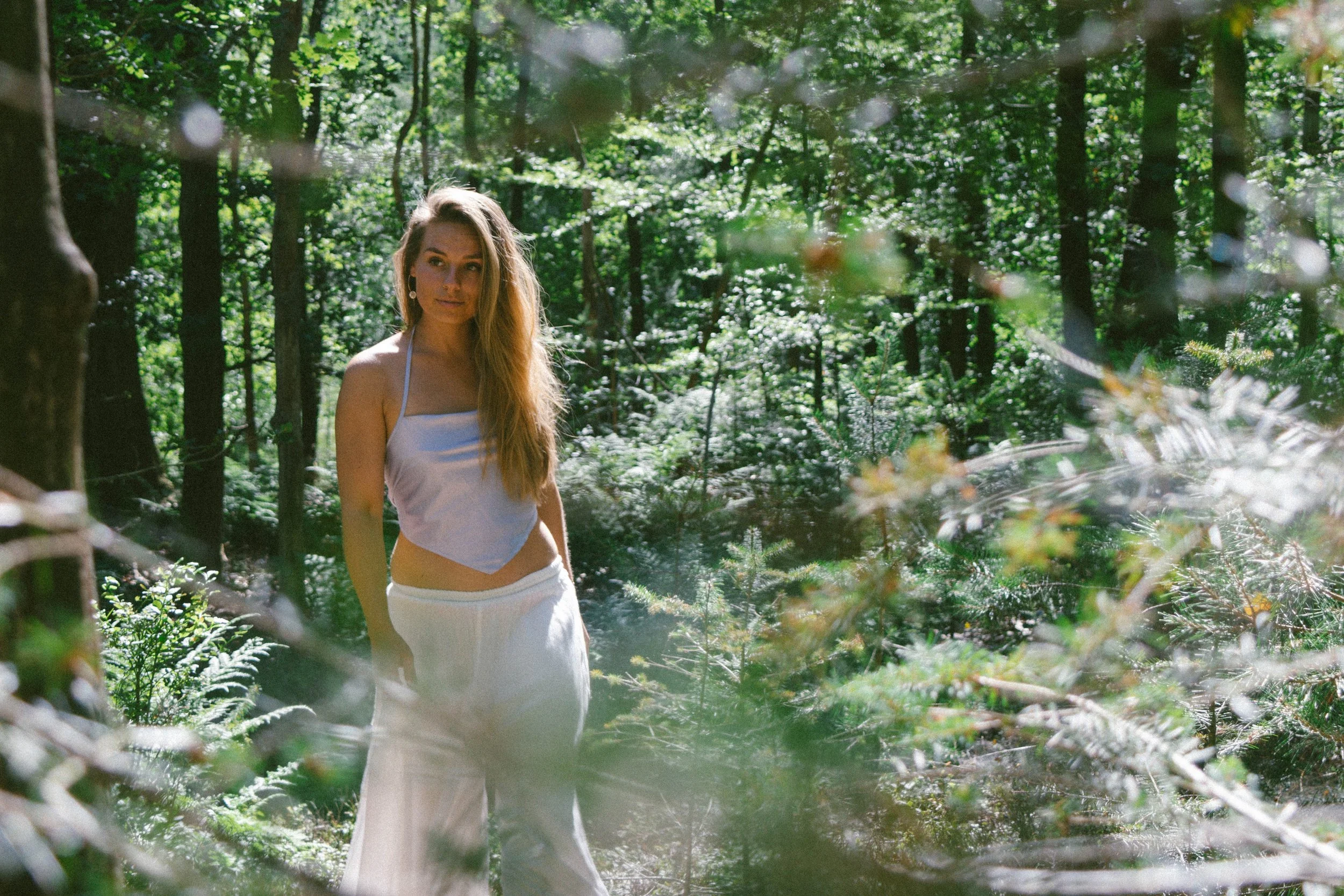 A young woman with long, wavy hair wearing a sleeveless white top and white pants stands in a lush, green forest with sunlight filtering through the trees.