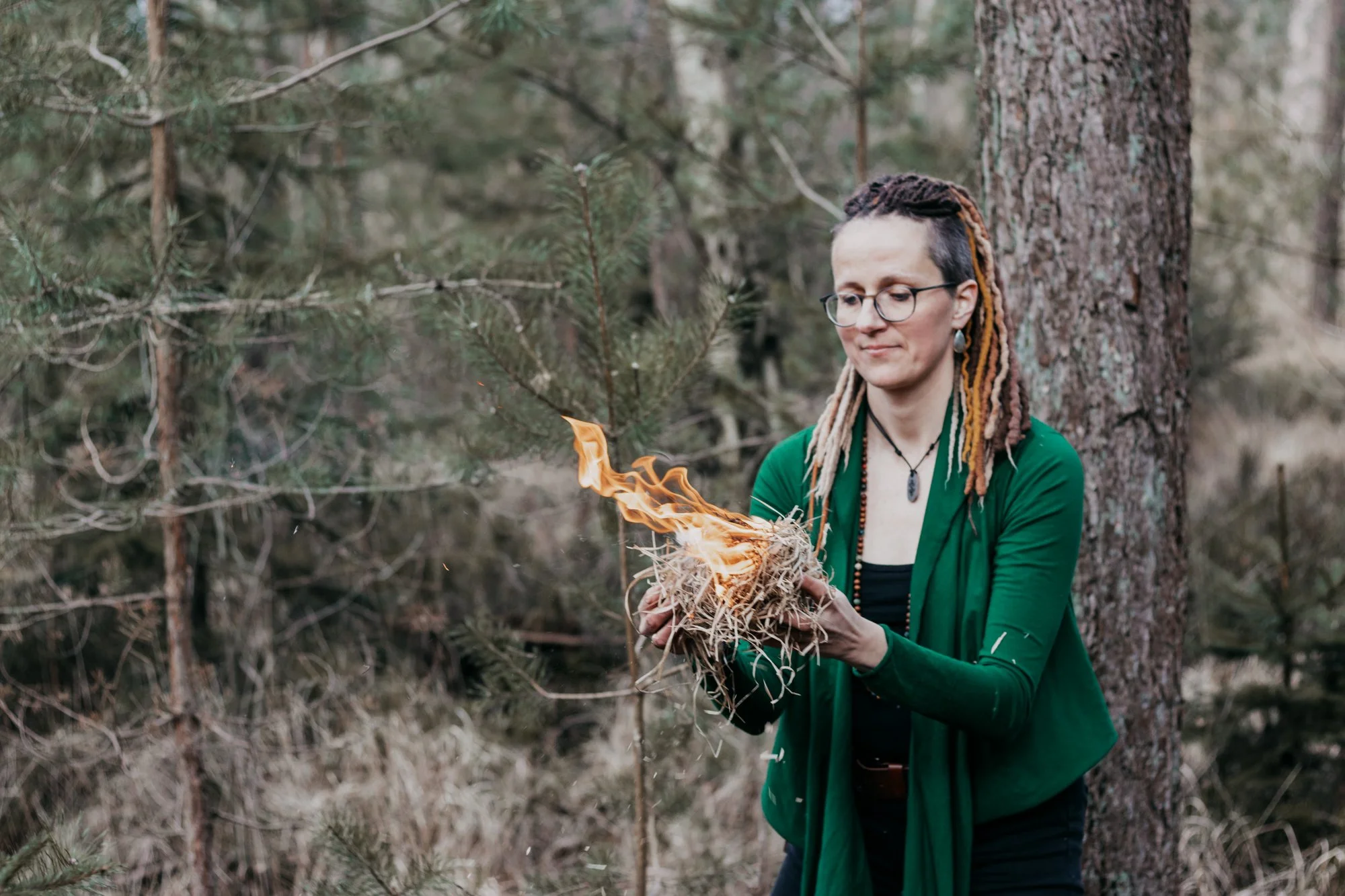 A woman with dreadlocks and glasses holding a burning bundle of dried twigs and leaves in a forest.