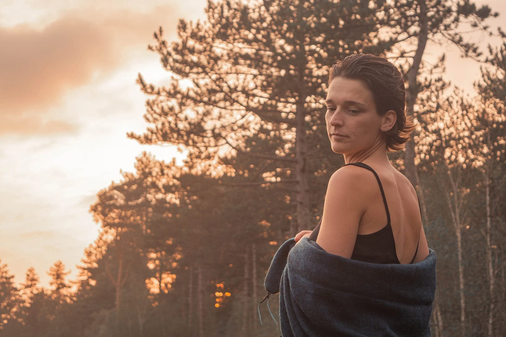 A woman with short brown hair wearing a black tank top and a dark sweater draped over her shoulders, standing outdoors during sunset with trees and a cloudy sky in the background.