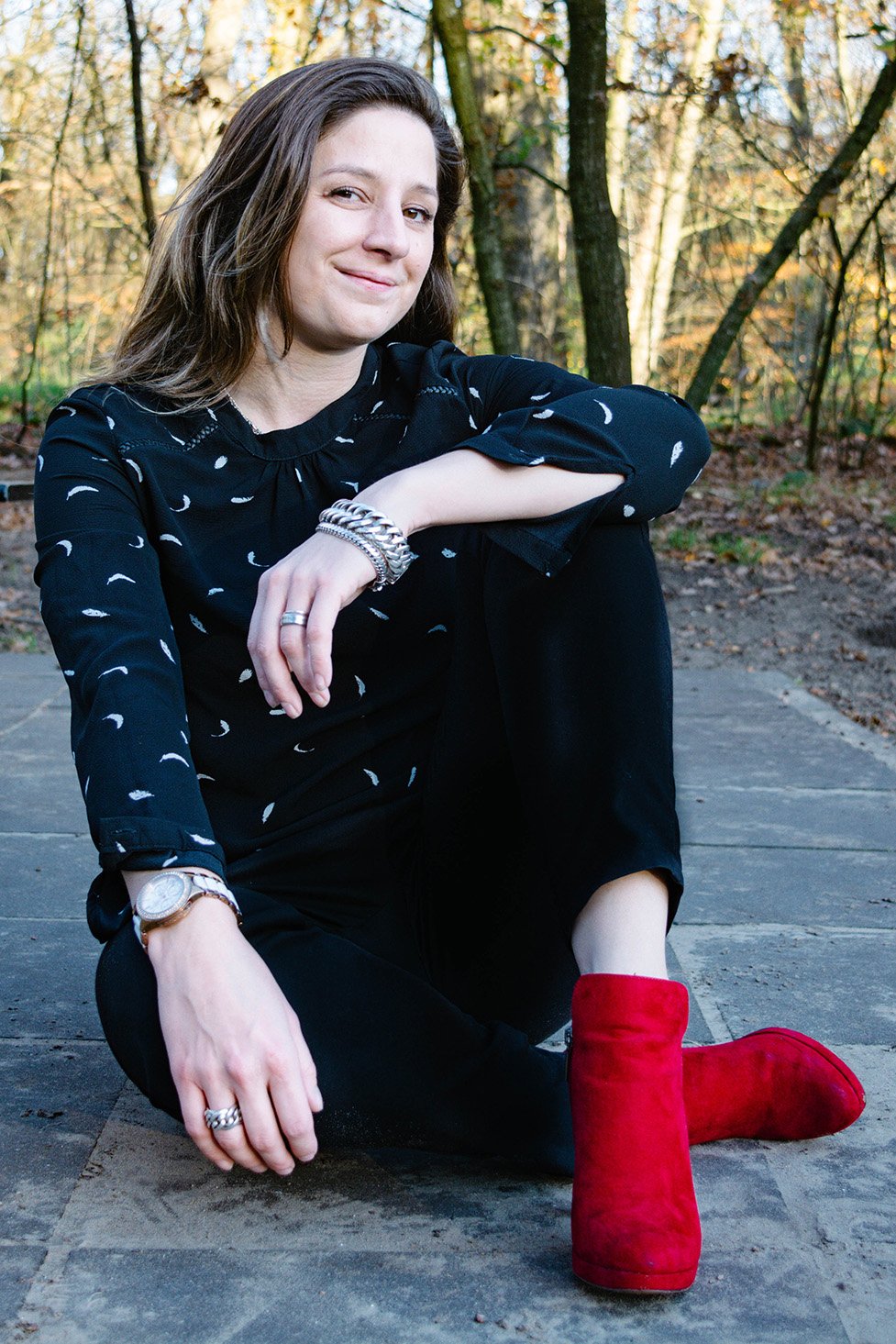 A woman sitting outdoors on a stone pathway during fall, wearing a black shirt with white patterns, black pants, red boots, and silver jewelry, smiling at the camera.