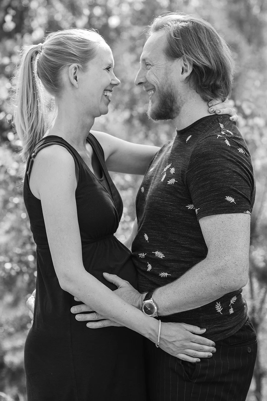A smiling pregnant woman with a ponytail and a sleeveless dress embraces a smiling man with shoulder-length hair and a beard, who is wearing a patterned T-shirt, outdoors with a blurred leafy background.