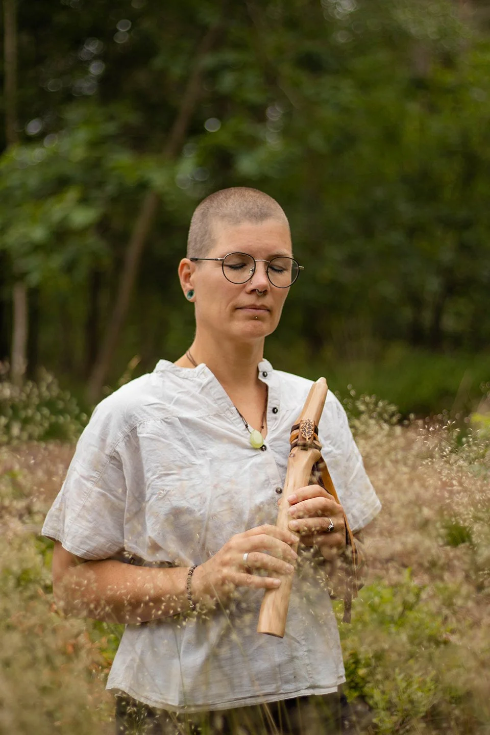 Person with short hair and glasses holding a wooden flute in a forested area.