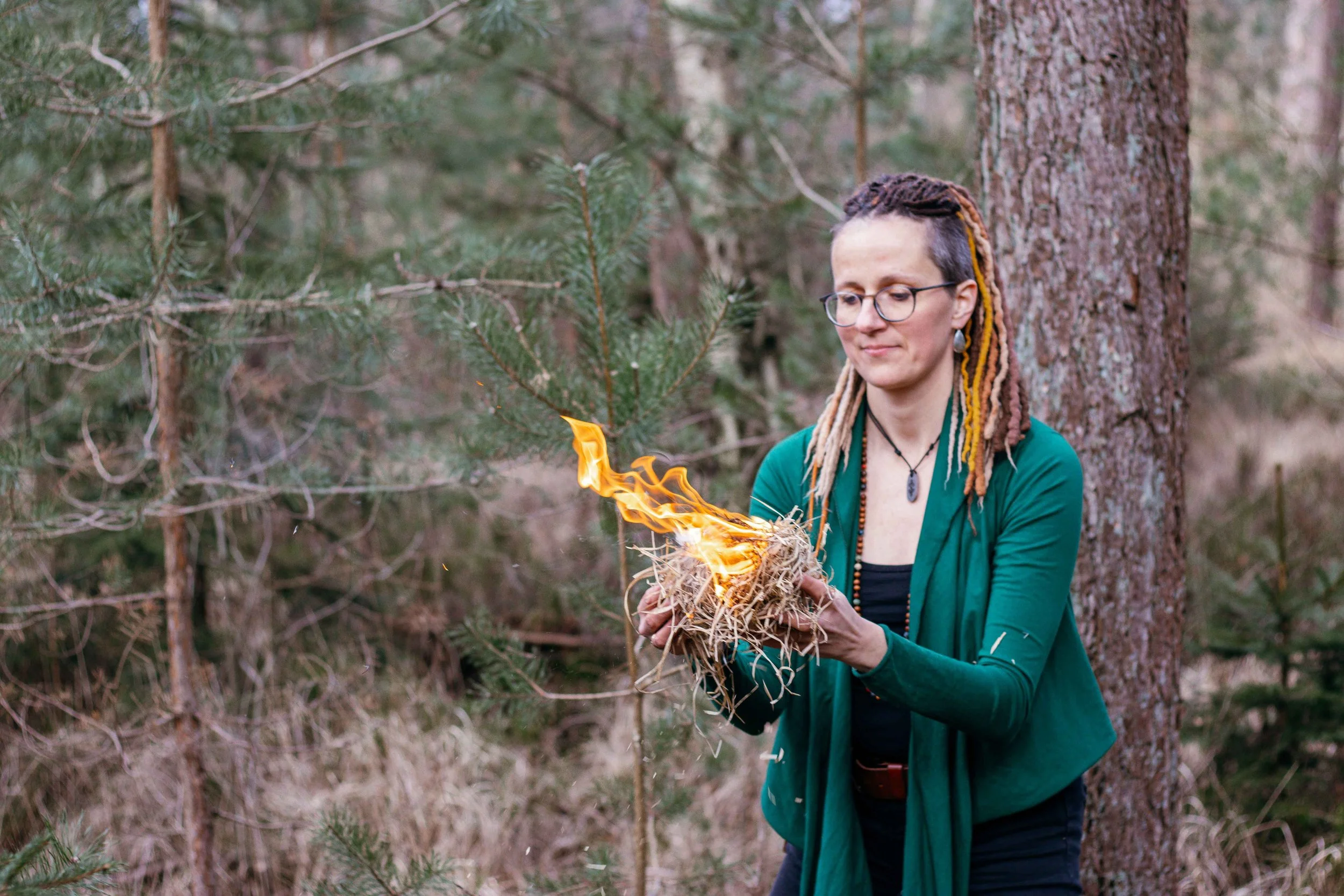 A woman in a green cardigan and glasses holding a small bundle of dry grass with flames on it in a forest