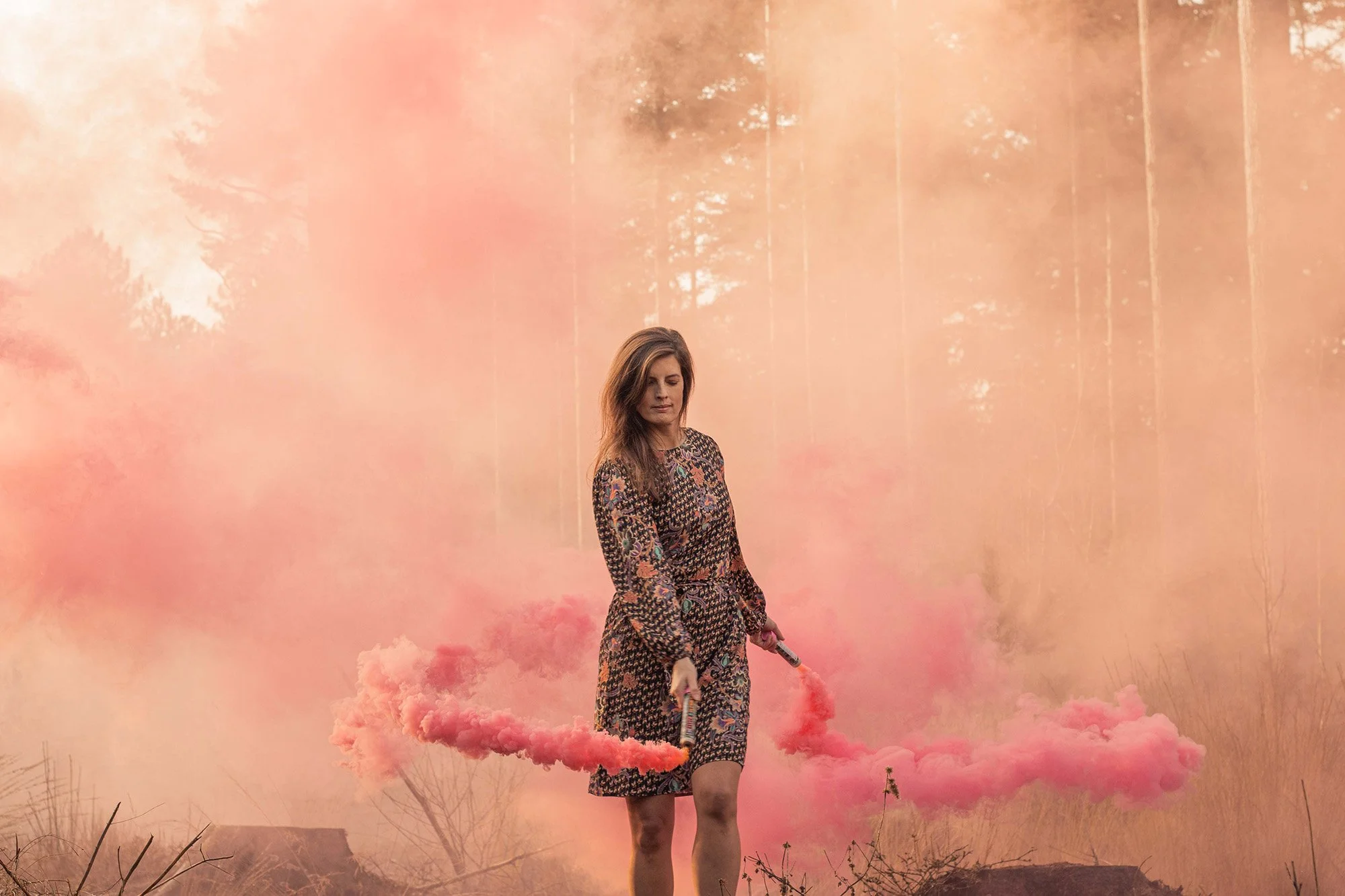 A woman in a patterned dress holding two pink smoke grenades, standing in a smoky forested area with trees in the background.