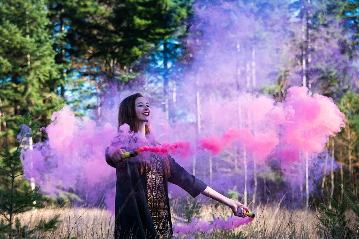 A woman smiling and holding two colorful smoke grenades, releasing pink and purple smoke in a forest setting with trees and blue sky